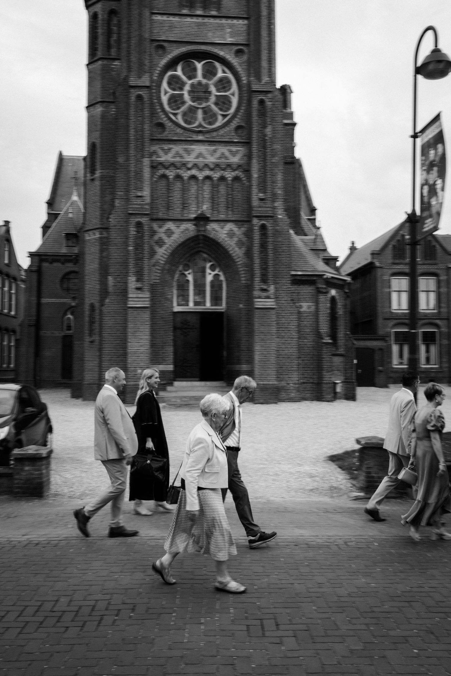 People walking on a brick plaza in front of a gothic church with a rose window in its tower on a rainy day.
