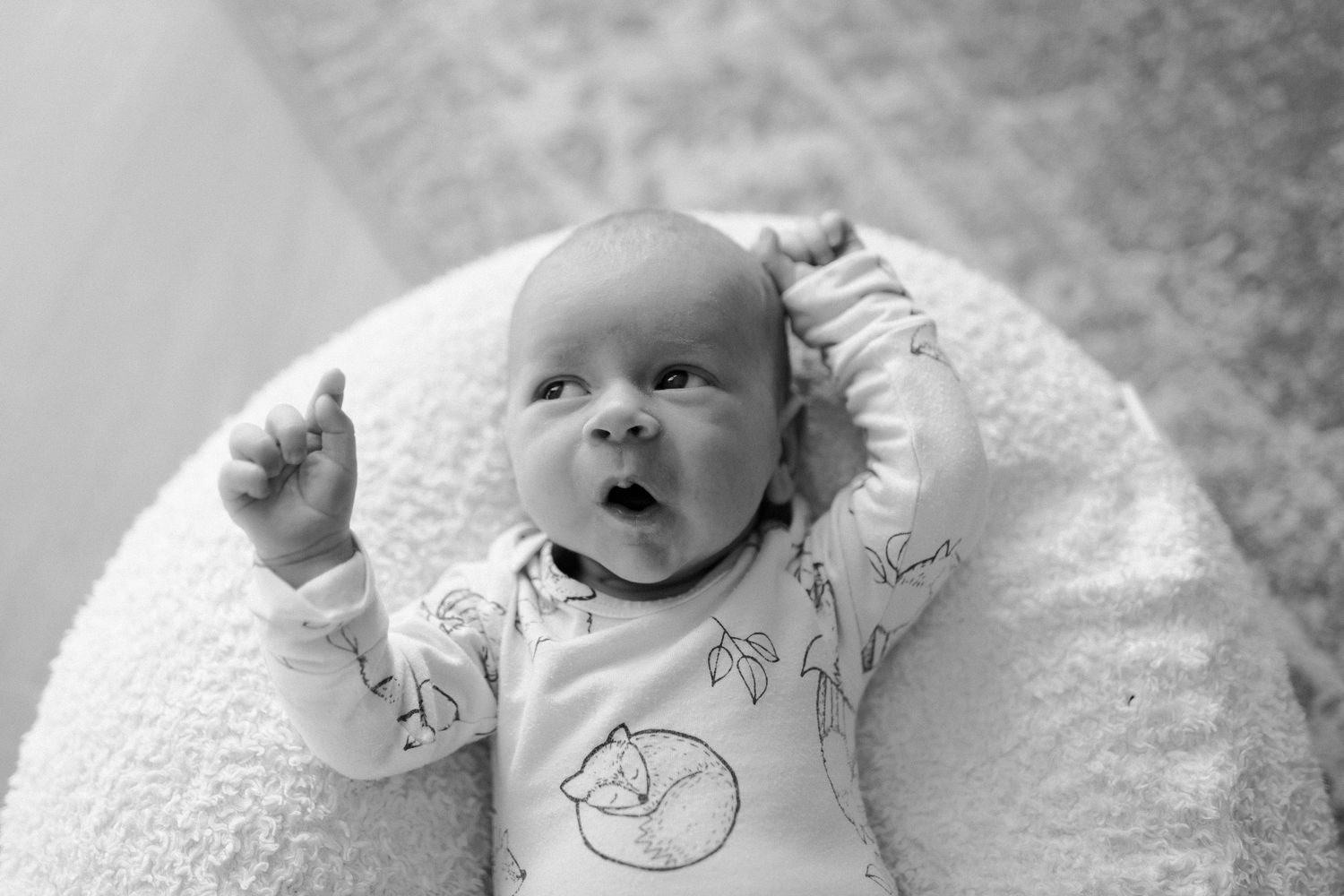 Black and white portrait of baby on round cushion with raised arms.