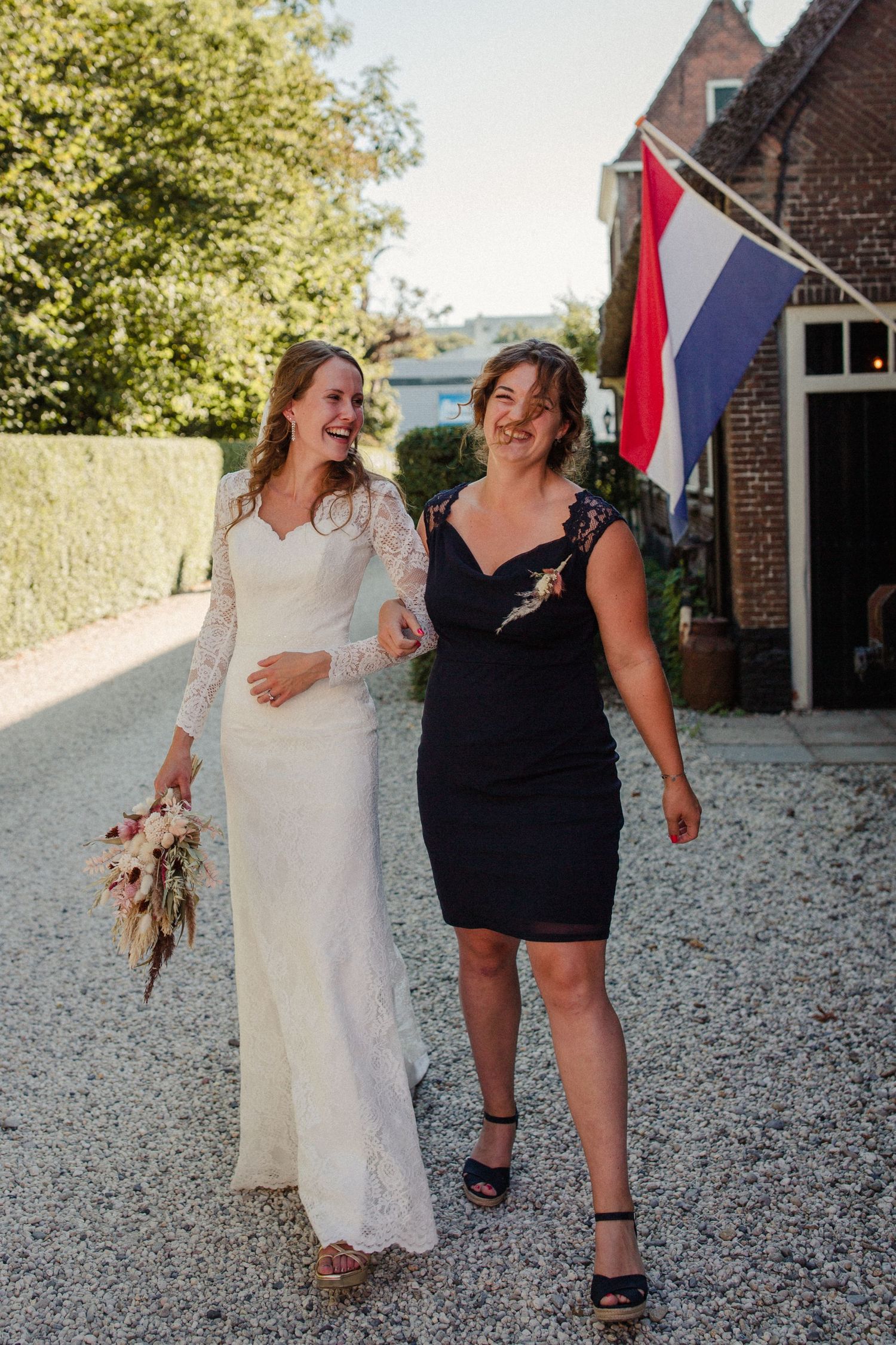 Two people walk and laugh together on a gravel path, with a Dutch flag visible in the background.