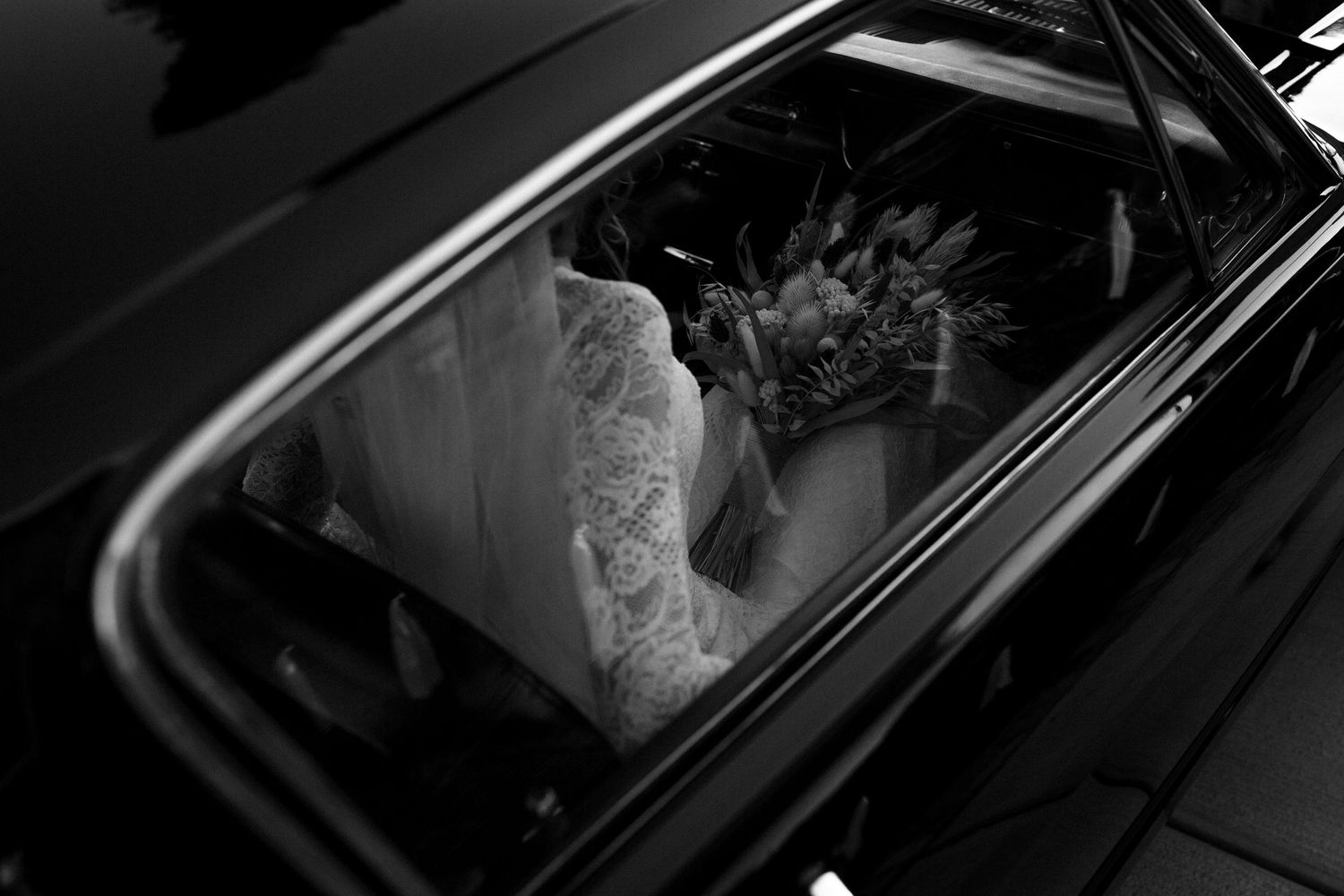 A black and white view through a car window showing someone in wedding attire sitting in the backseat.