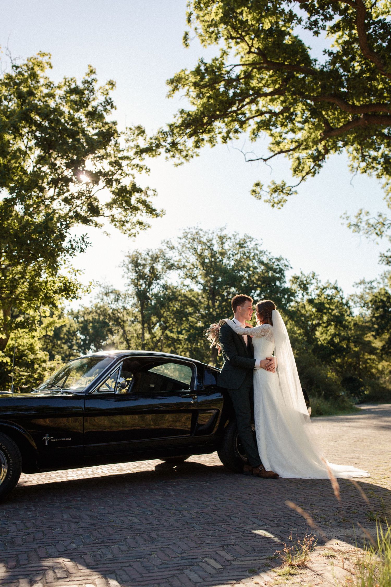 A romantic couple poses beside a vintage black car on a brick pathway surrounded by sunlit trees.