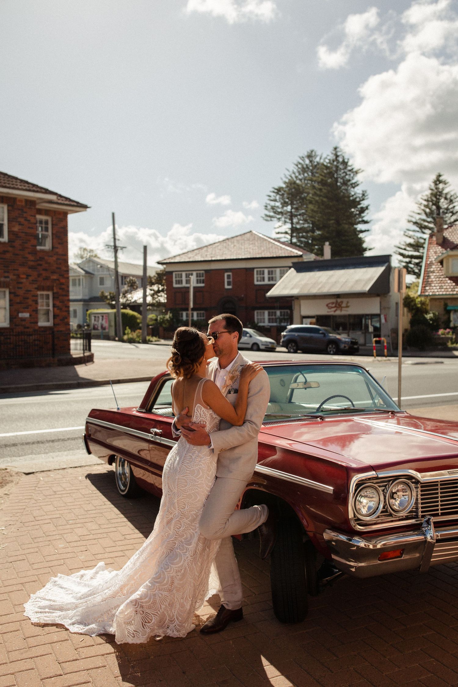 A romantic wedding couple poses with a vintage red car outside brick buildings.
