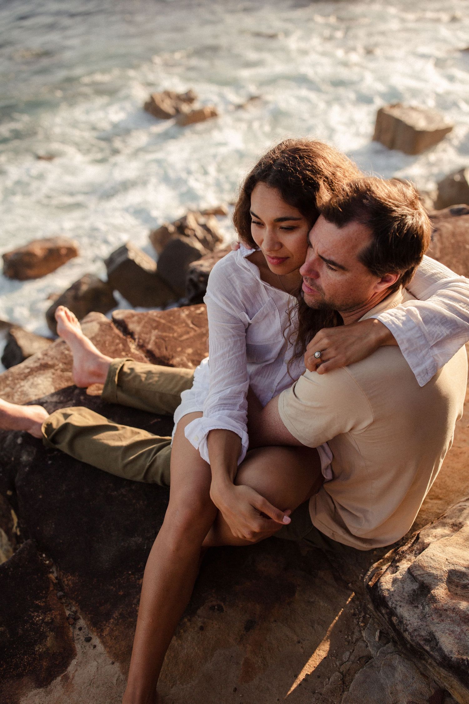 A romantic couple embraces while sitting on coastal rocks during golden hour, creating an intimate moment.