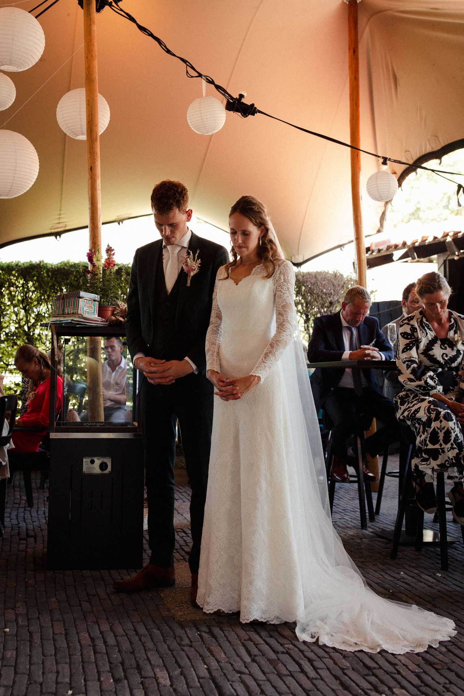 A couple stands under a tent decorated with white lanterns at an outdoor wedding ceremony.