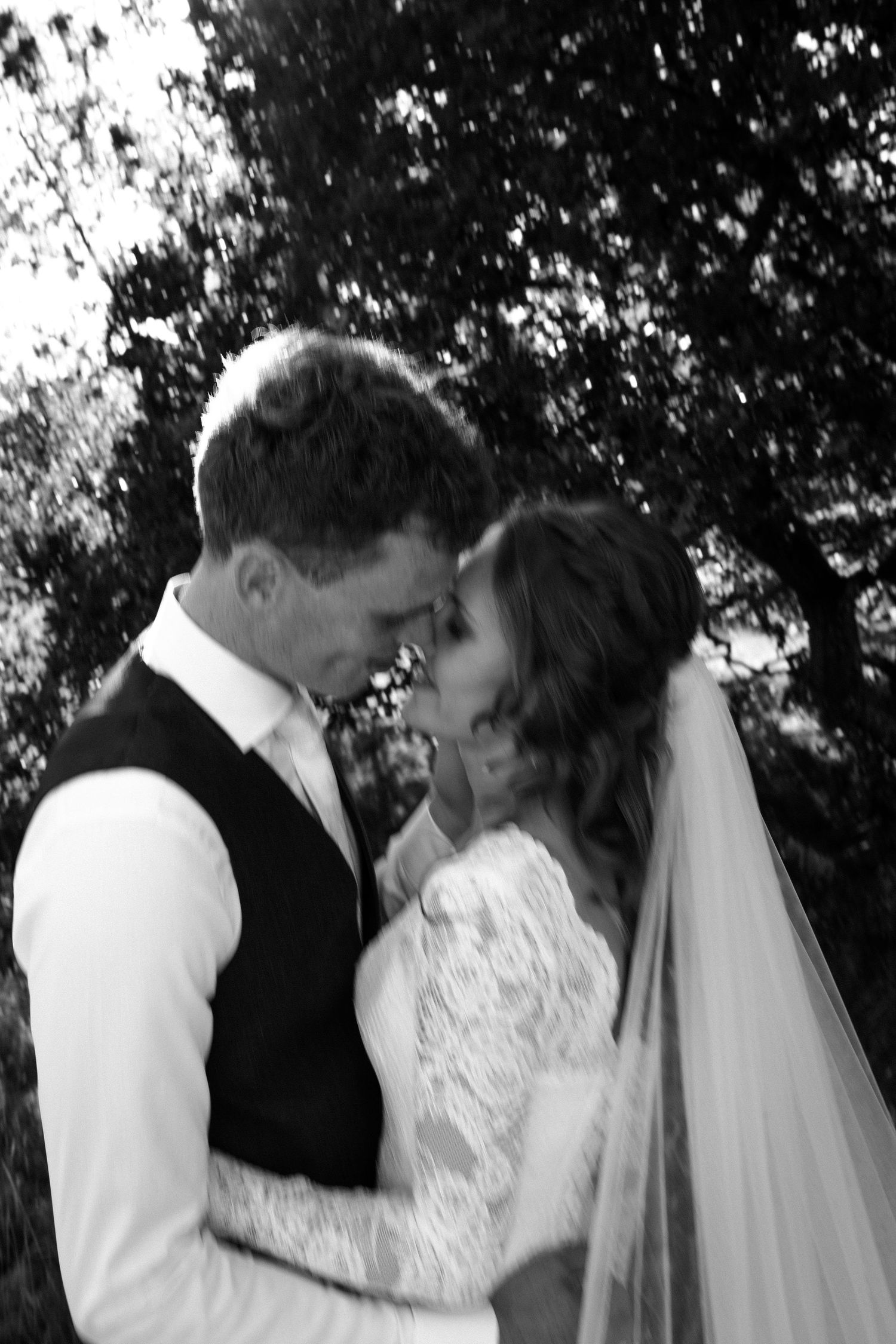 A tender moment between newlyweds in formal wedding attire sharing an intimate moment outdoors in black and white.