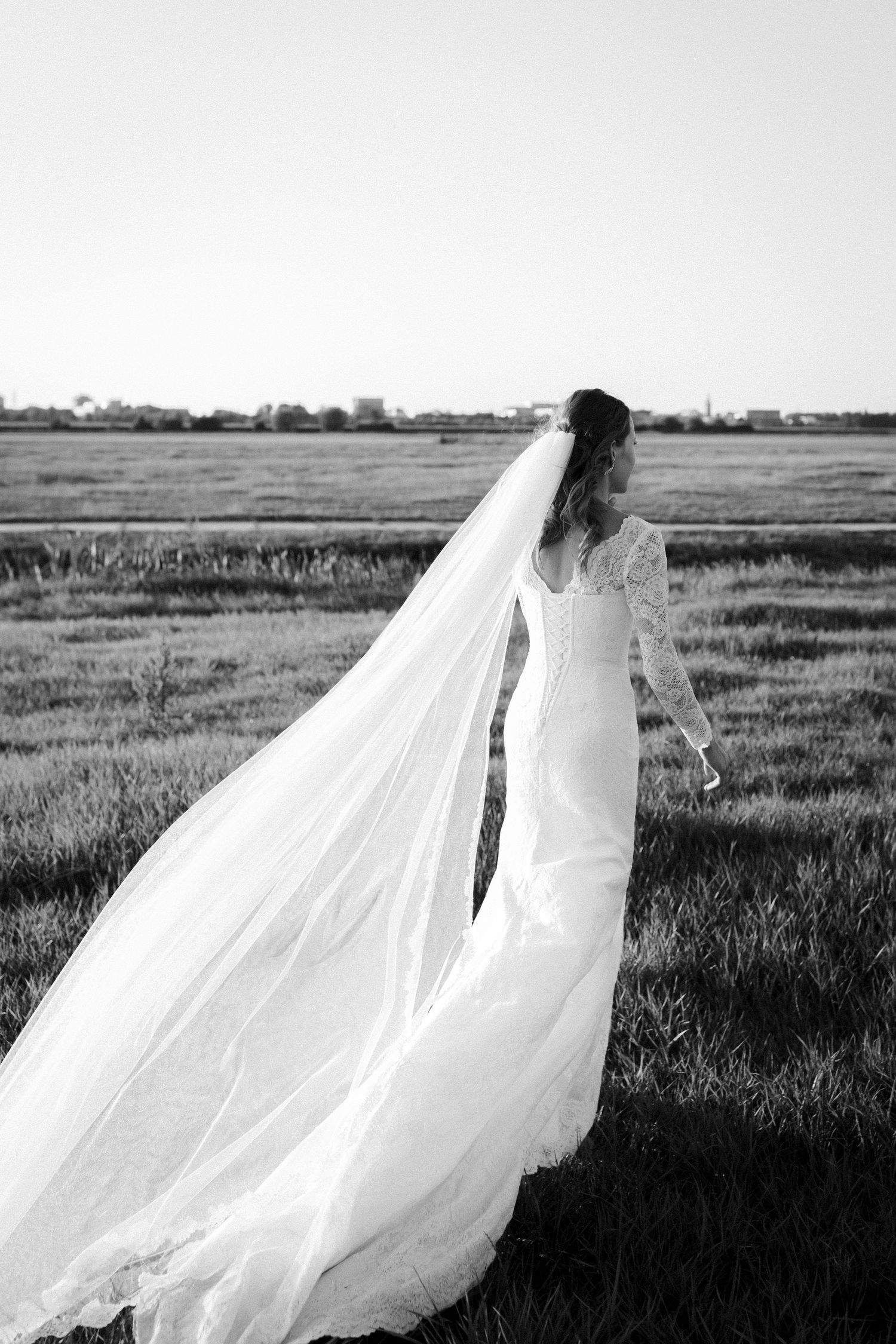 A bride in a lace long-sleeve wedding gown with flowing veil stands in a grassy field at dusk.