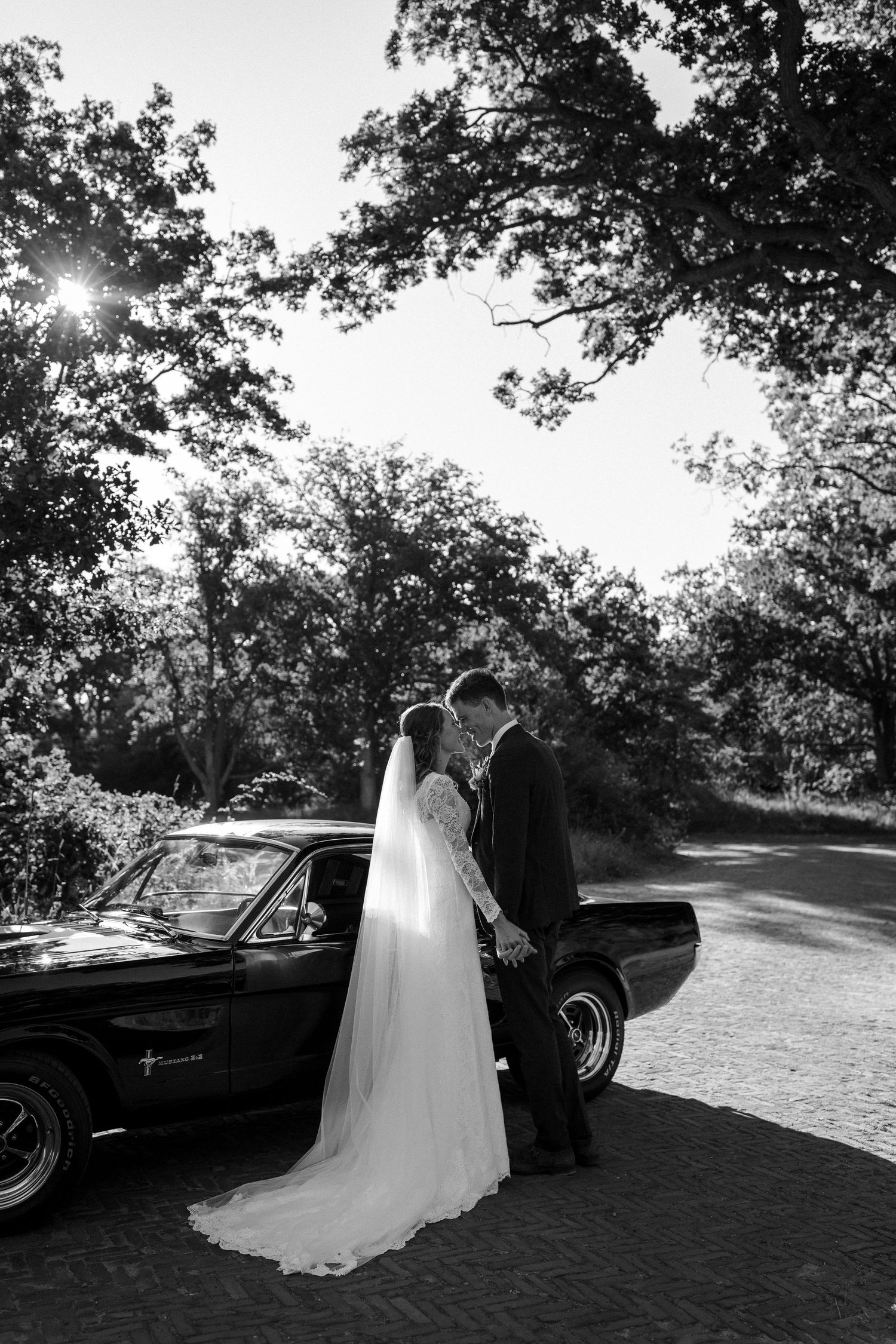 Black and white wedding photo with a couple standing by a classic car beneath sun-dappled trees on a winding road.