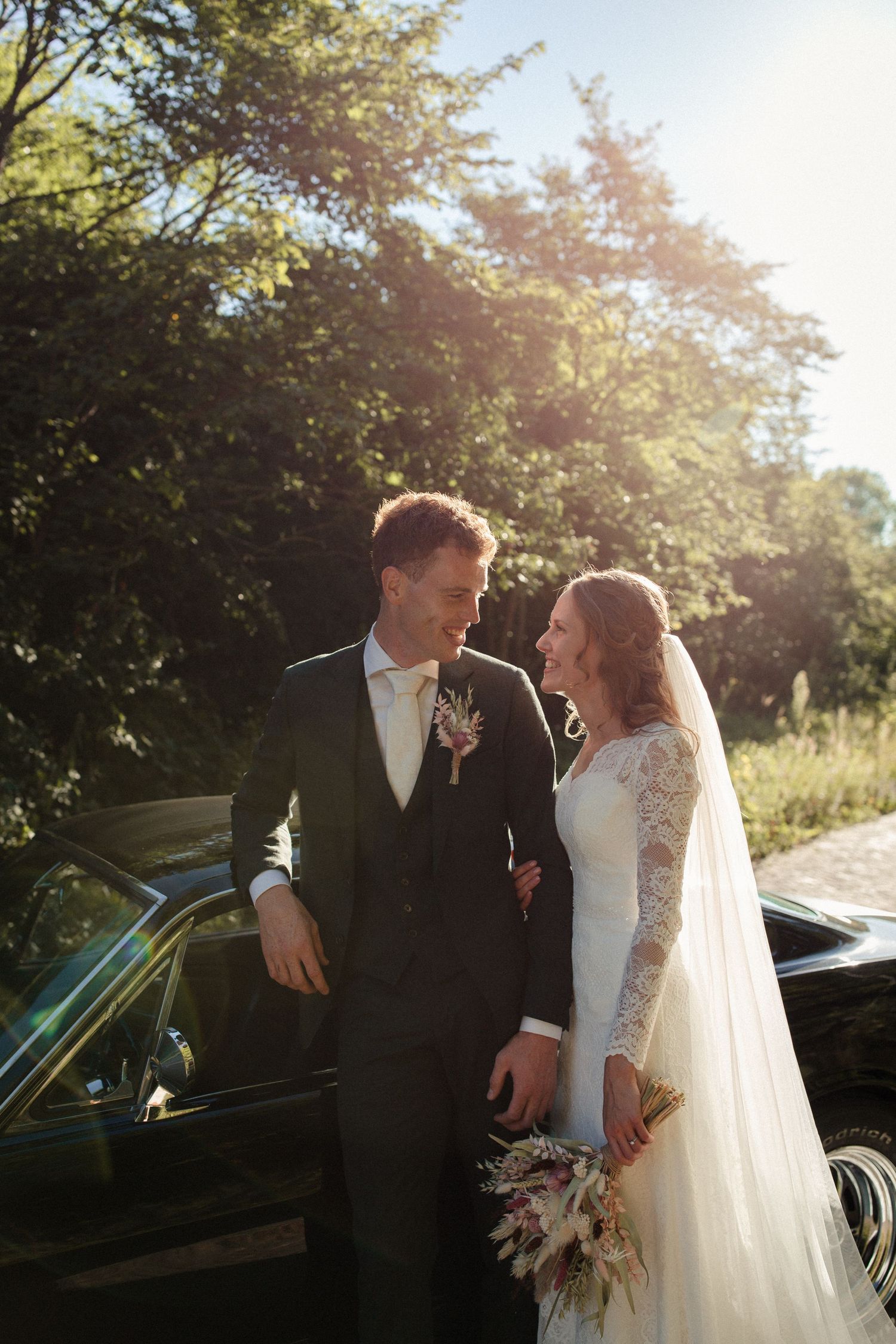 A newlywed couple sharing a joyful moment beside a vintage car on their wedding day in golden sunlight.