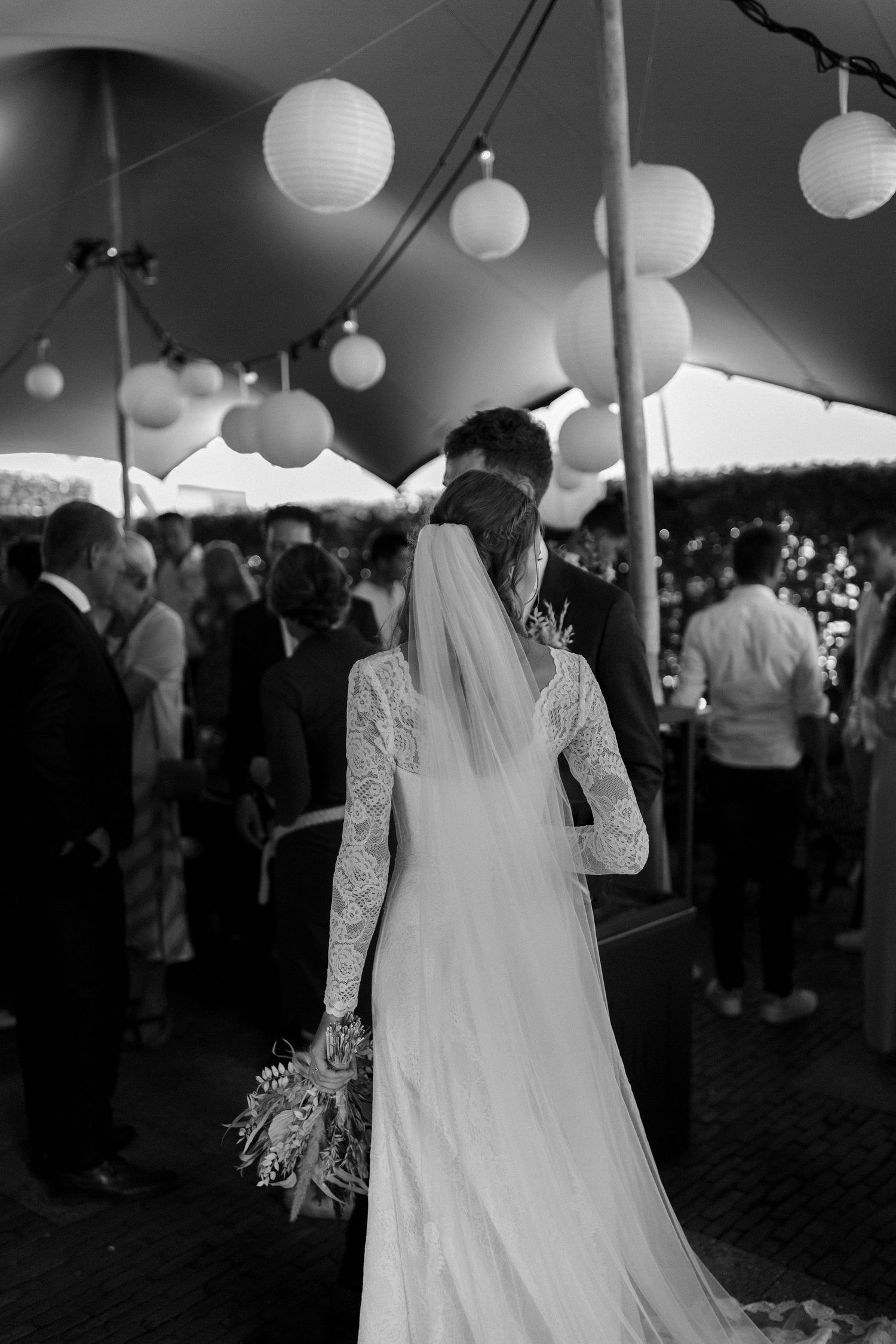 A bride in a long-sleeved lace wedding dress stands under white paper lanterns at an outdoor reception tent.