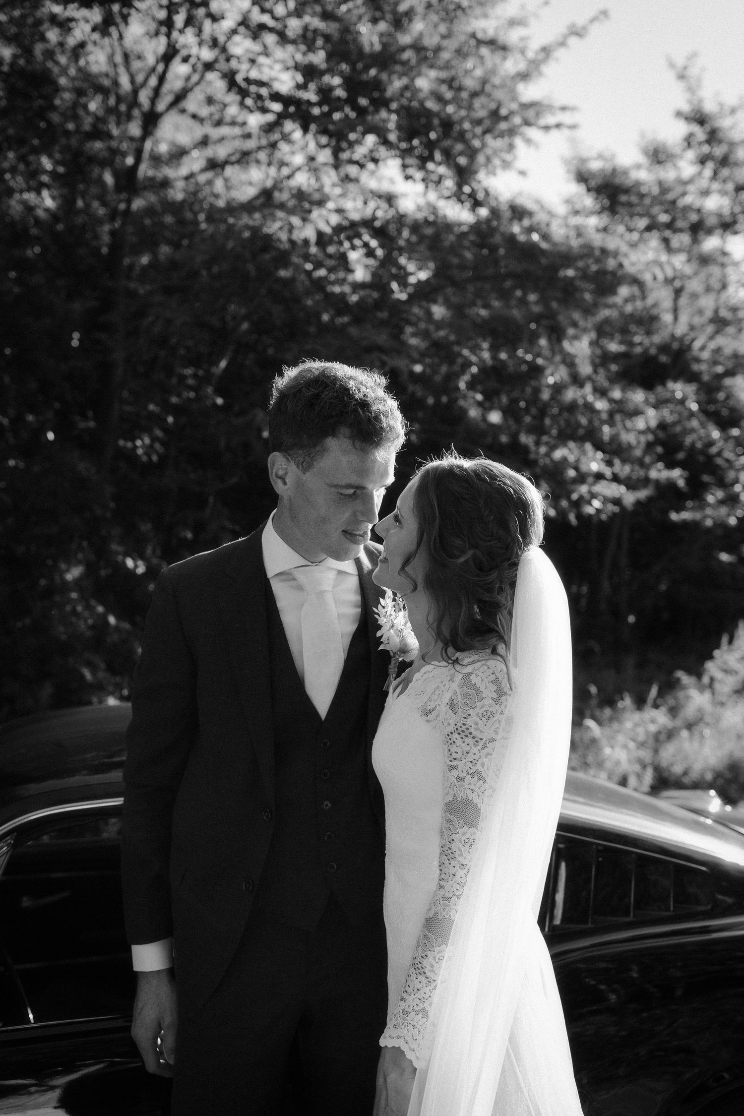 Romantic black and white wedding photograph of newlyweds sharing an intimate moment next to a car outdoors.