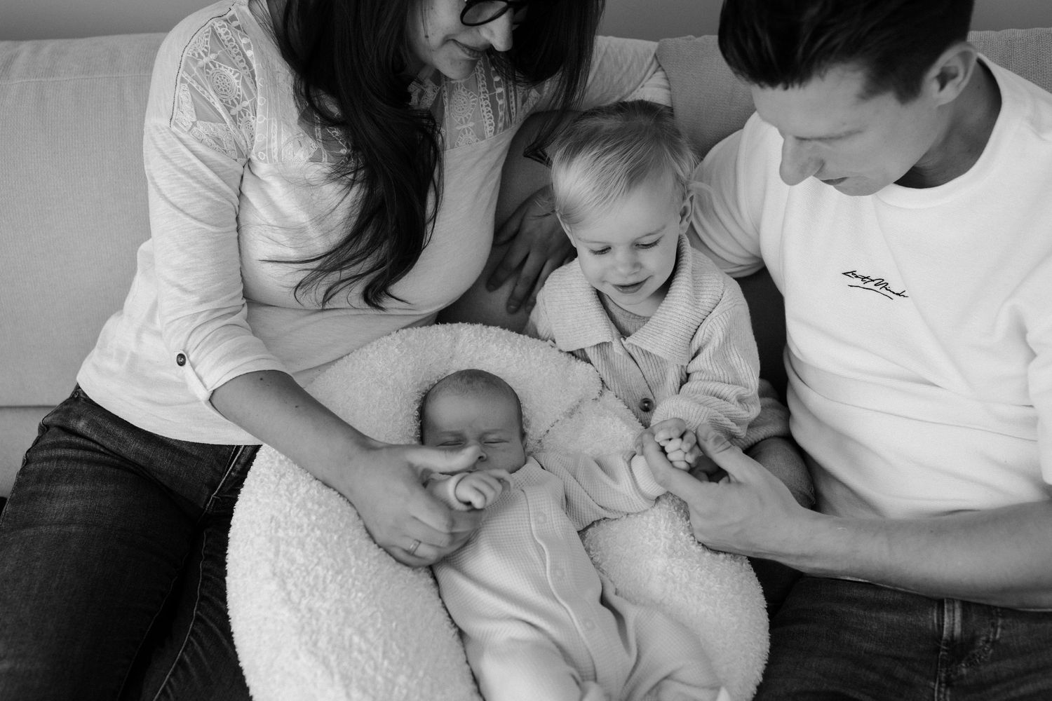 Black and white intimate family photo of parents and toddler sitting together on white couch.