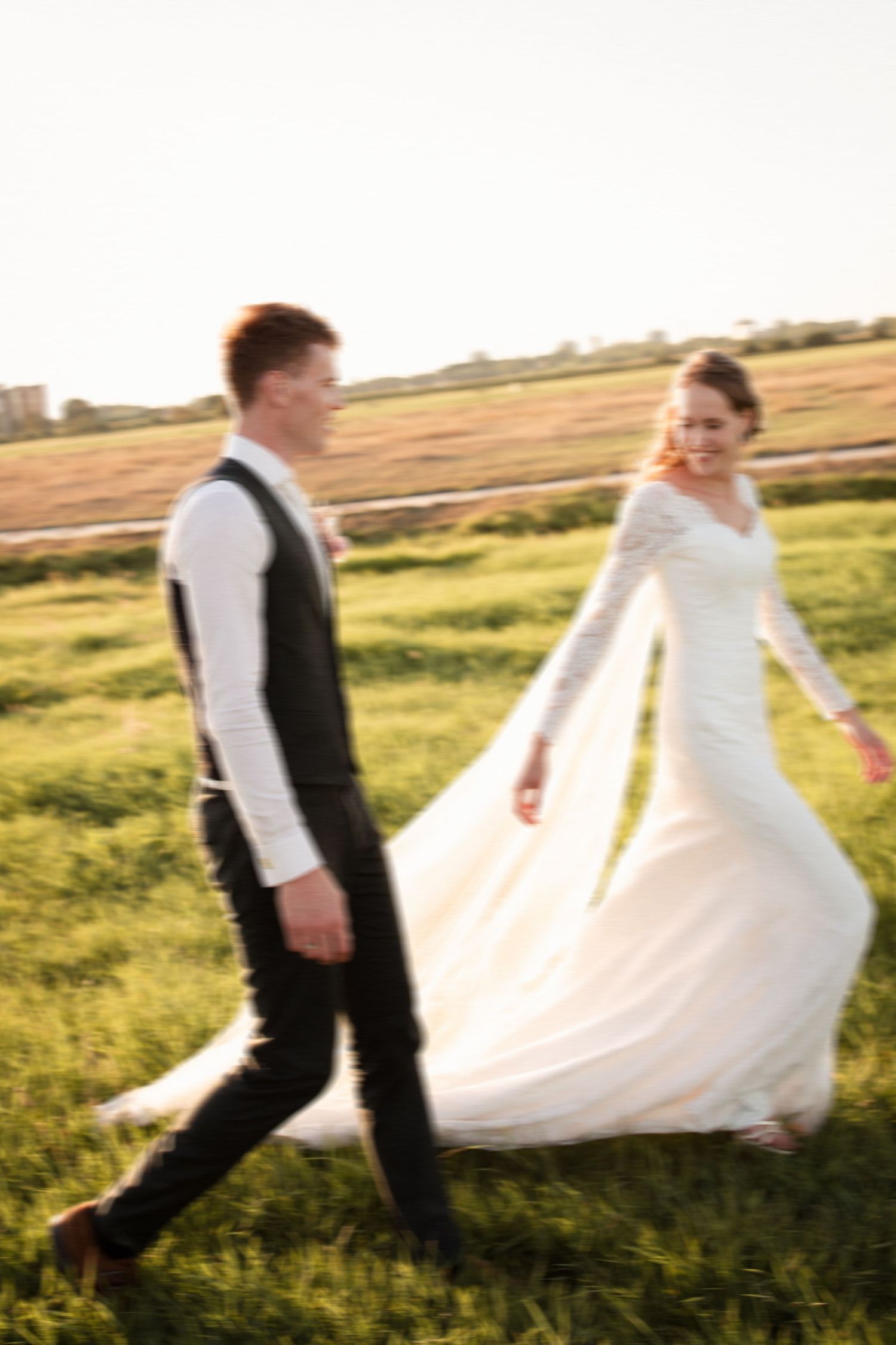 A couple walks through a sunlit grassy field at sunset, with the bride's white dress flowing in the breeze.