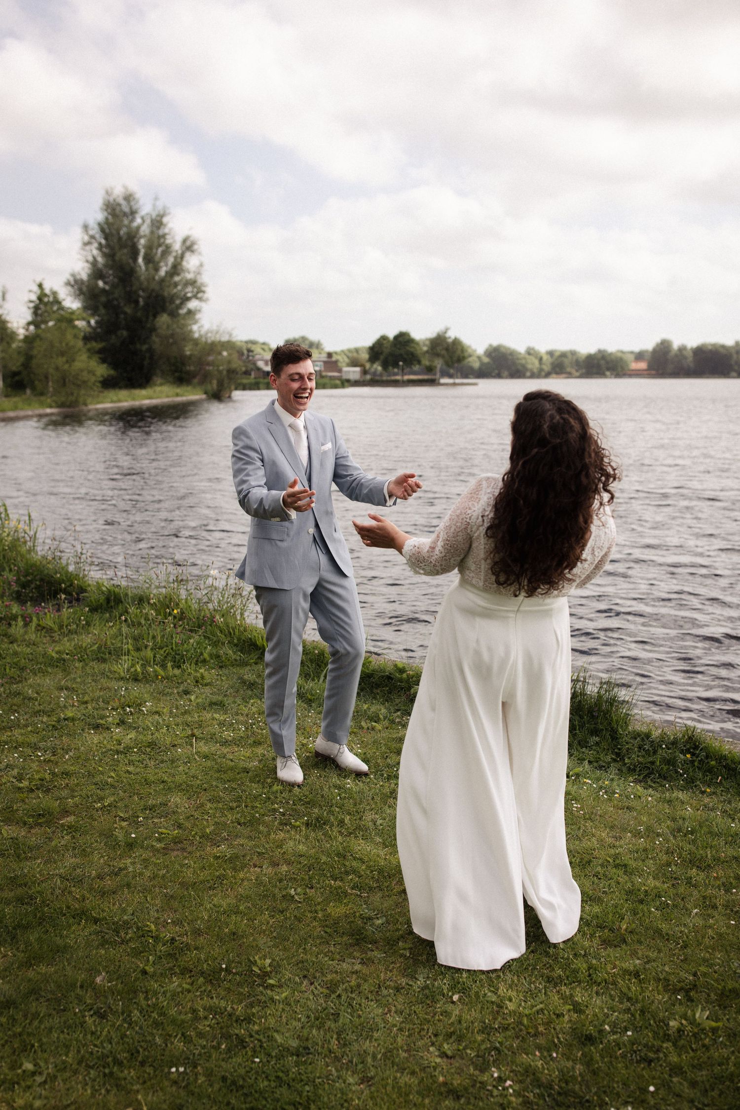 A couple in wedding attire playfully interacts on a grassy shore beside a scenic lake.