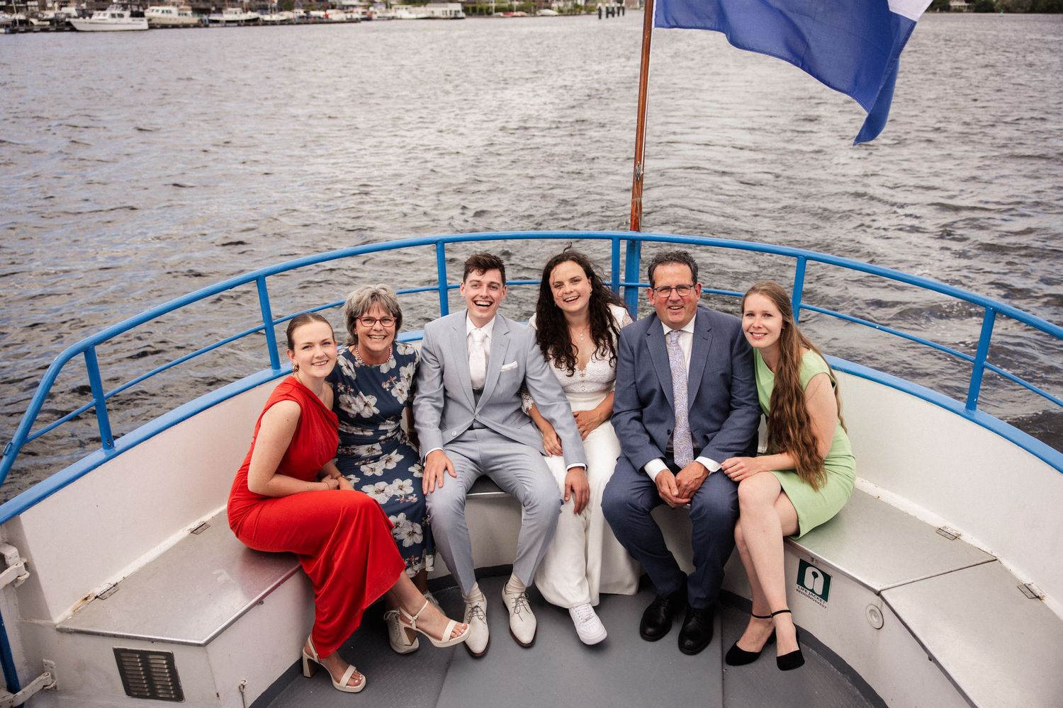A group of well-dressed guests sitting together on a boat deck with a blue flag waving behind them.