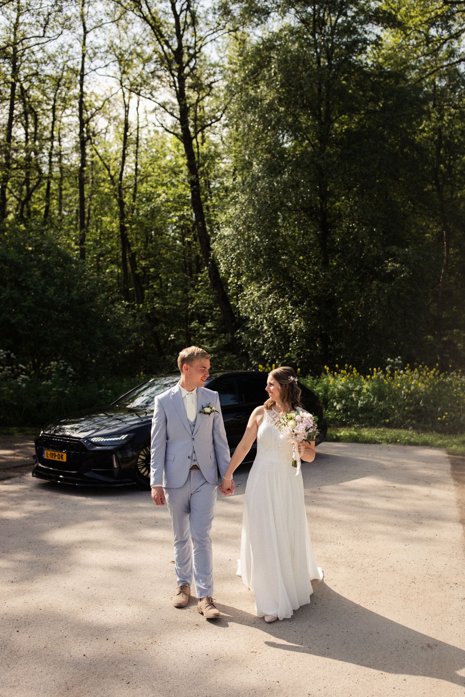 Newlyweds walk hand in hand on a sun-dappled path with their car parked behind them in a wooded setting.