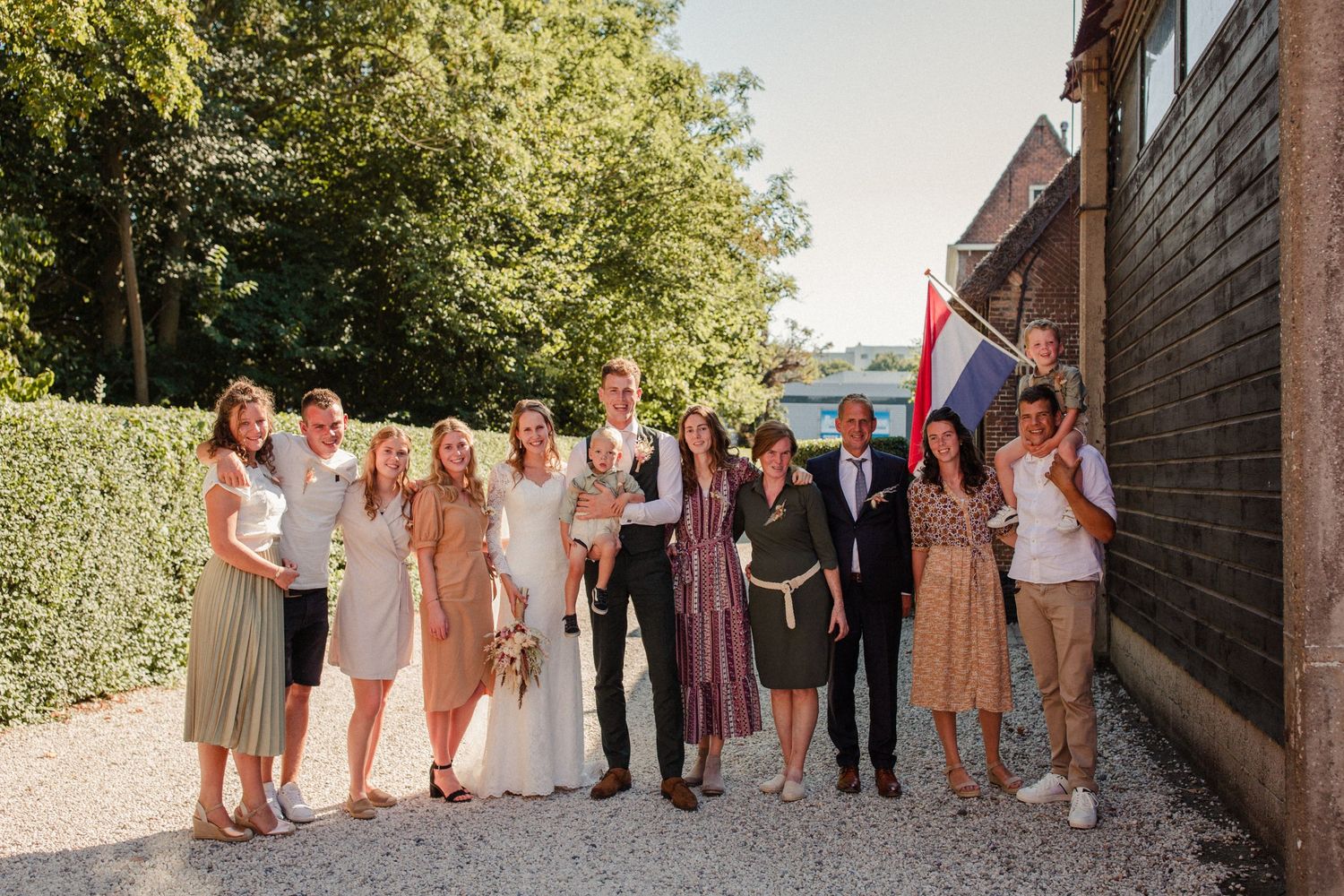 A group of stylishly dressed wedding guests stand together on a gravel path next to a rustic building on a sunny day.