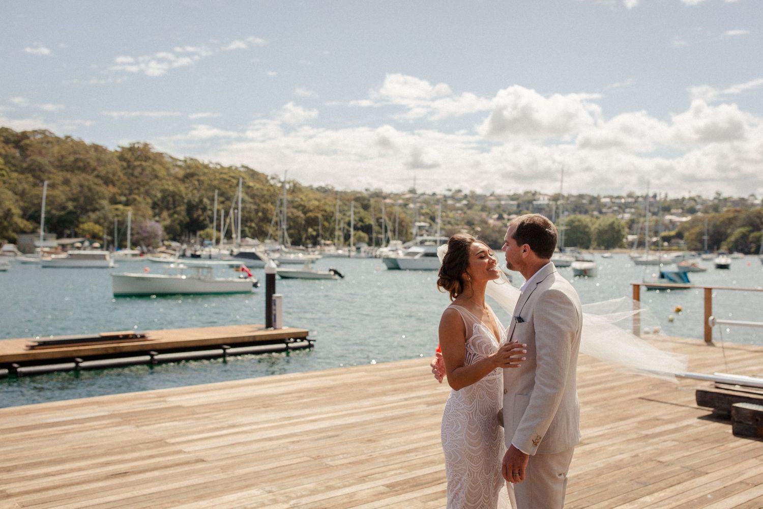 Couple in wedding attire embrace on waterfront dock with boats in background.