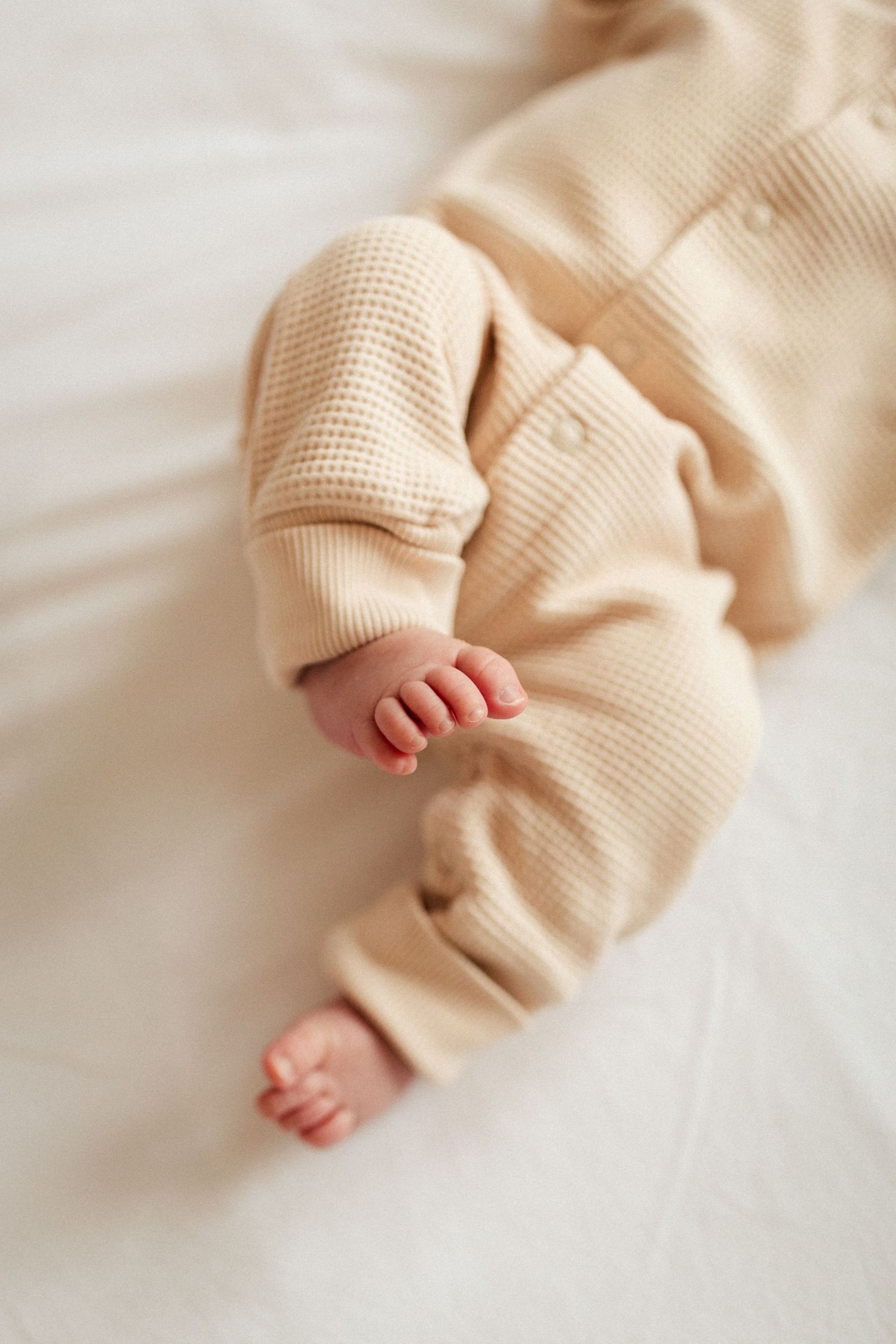 Tiny baby feet peeking out from cozy beige knit clothing against white bedding.