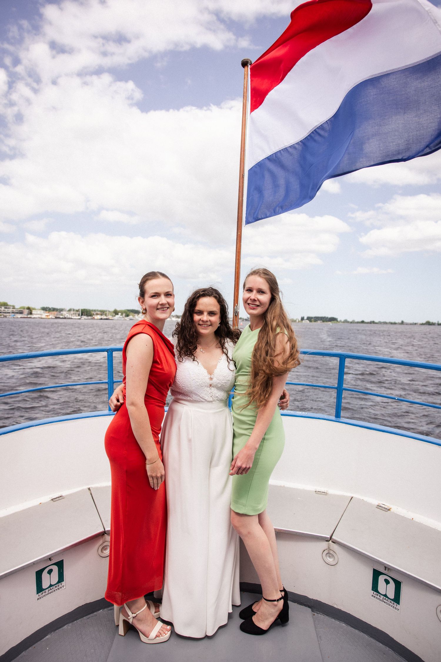 Three people in formal dresses pose on a boat deck with a Dutch flag flying overhead on the water.