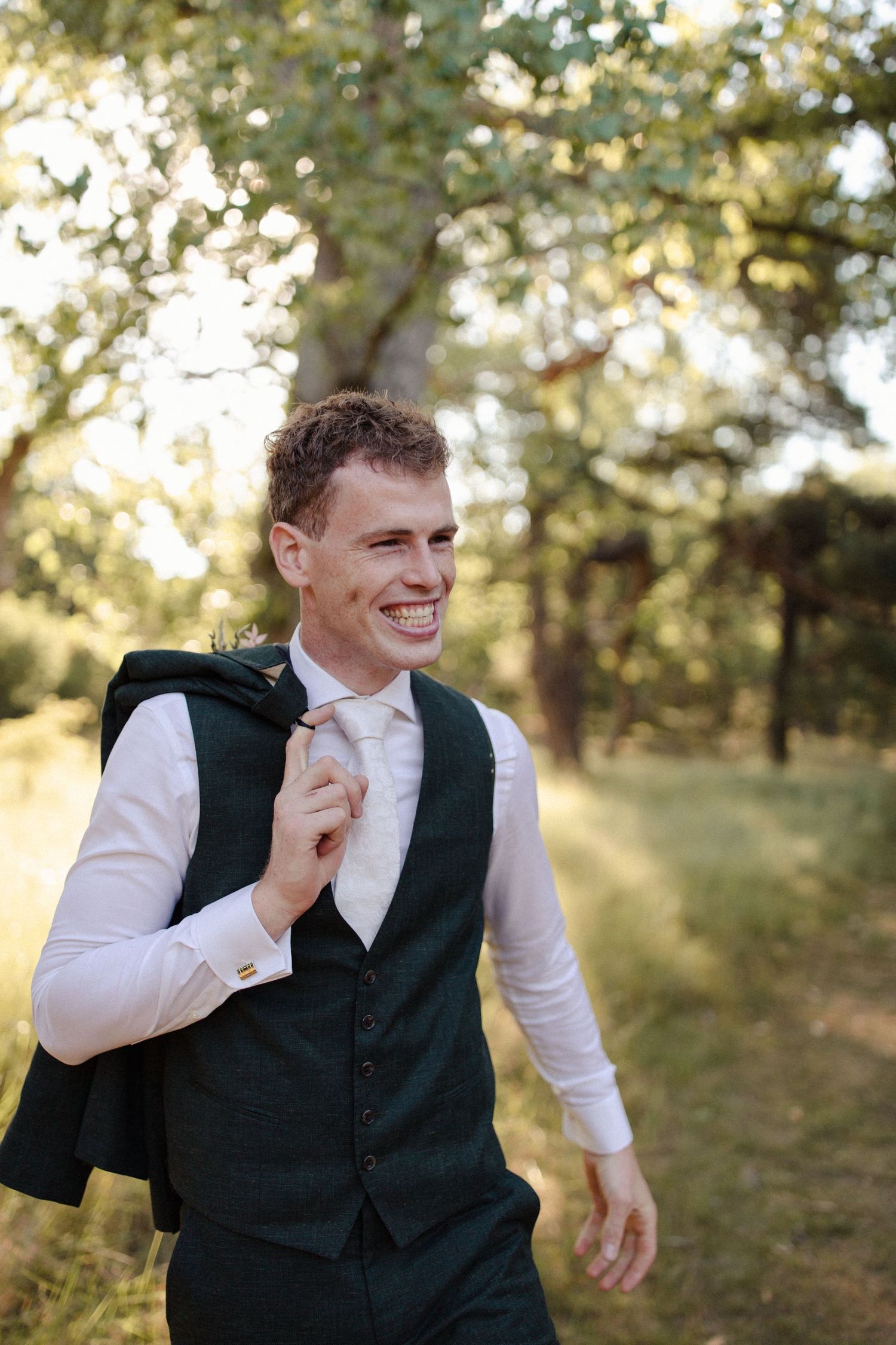 A person wearing a formal vest and white dress shirt smiles joyfully in a sun-dappled outdoor setting.