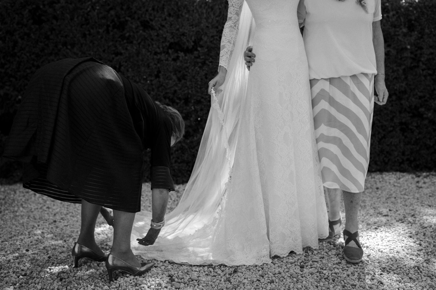 Black and white photo showing details of a wedding dress hem and shoes on a gravel path during a wedding ceremony.