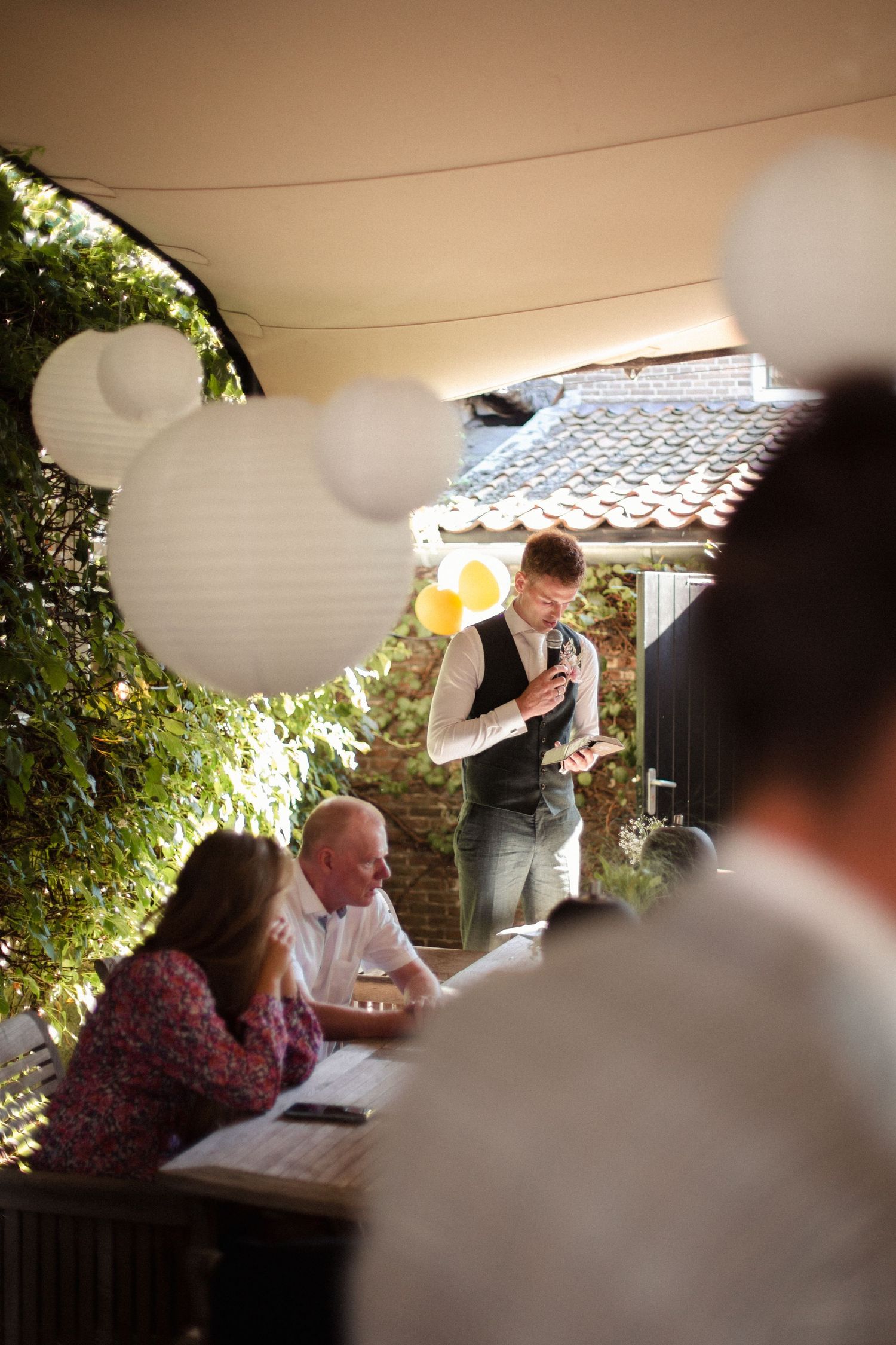 Wedding reception guests gather under white paper lanterns at an outdoor venue with tiled roofing.