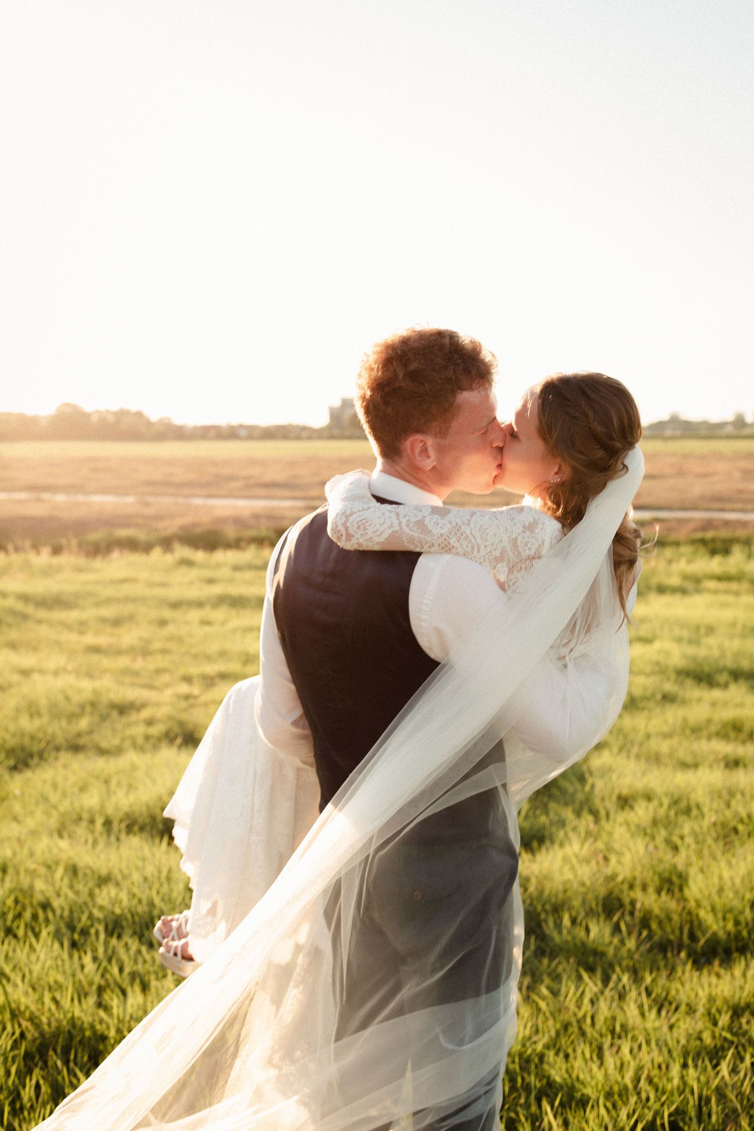A romantic wedding couple shares a kiss in a sunlit field at golden hour as the bride's veil flows in the breeze.