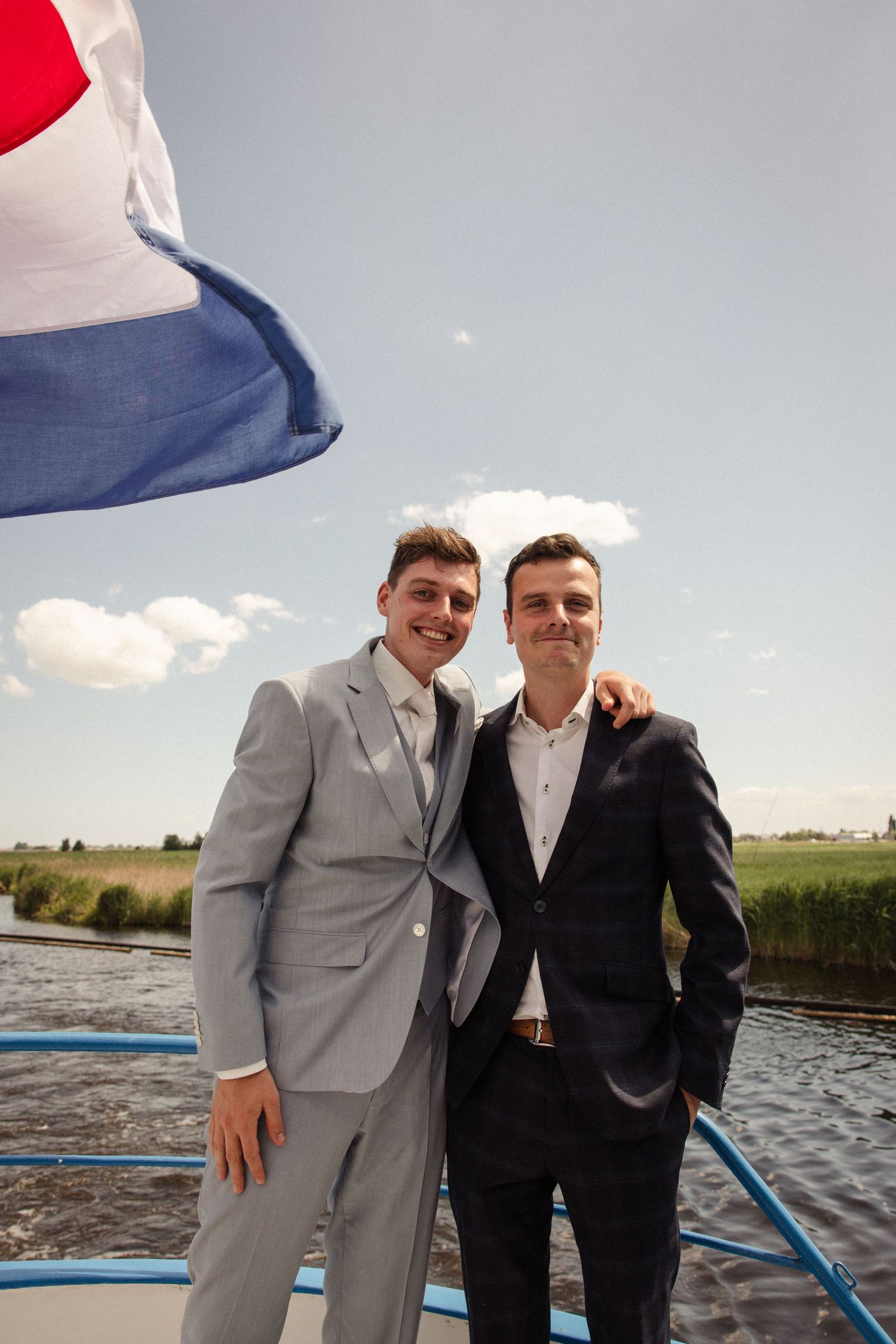 Two people in formal attire stand on a boat deck against a scenic waterway backdrop on a sunny day.