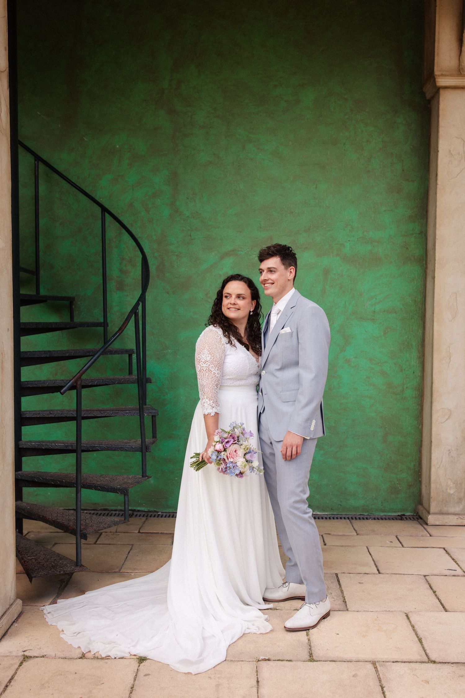 Wedding portrait of couple against green wall with spiral staircase, bride holding purple bouquet.