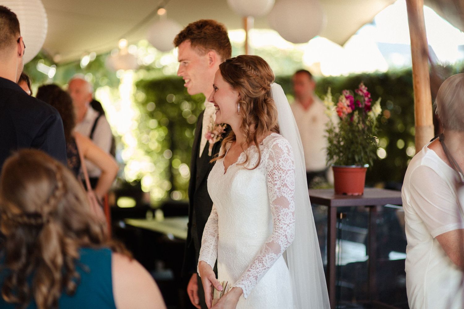 The bride in a long sleeve lace wedding dress smiles during an outdoor reception under a white tent with floral decor.