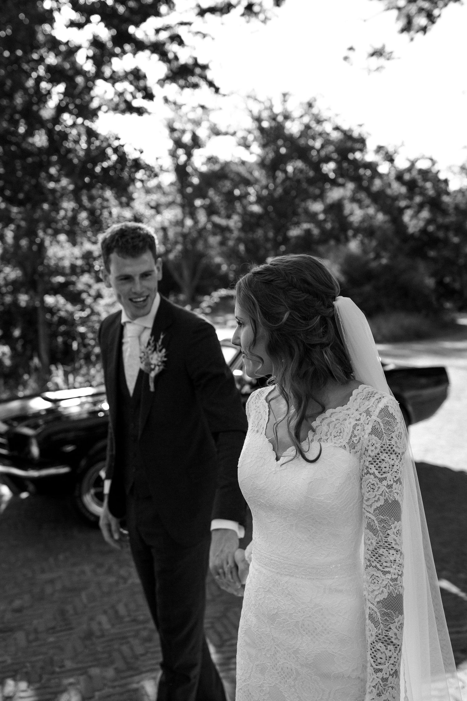 Black and white photo of a bride in a lace-sleeved gown and flowing veil walking outdoors with her groom on wedding day.