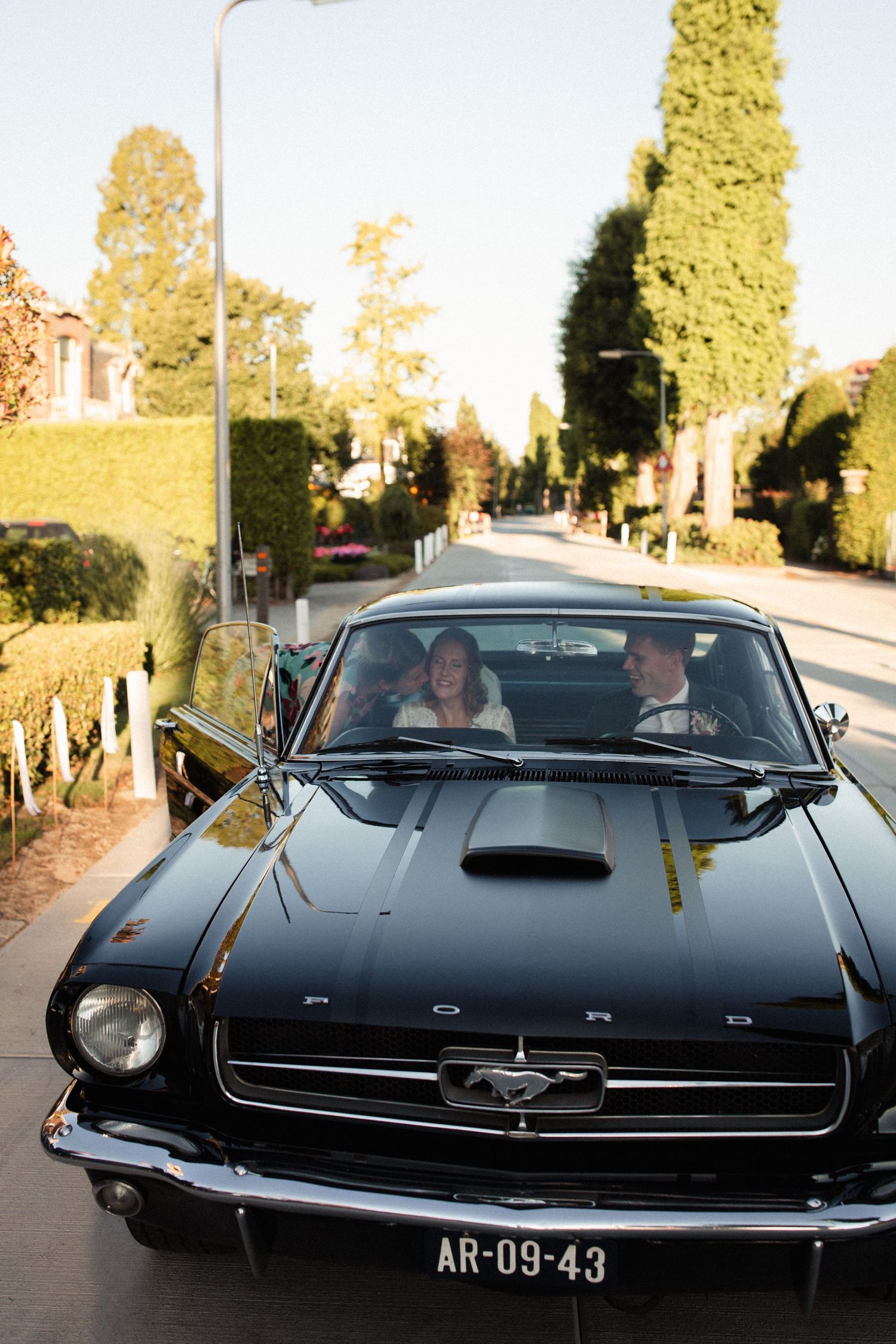 A classic black Ford Mustang convertible driving down a tree-lined residential street on a sunny day.