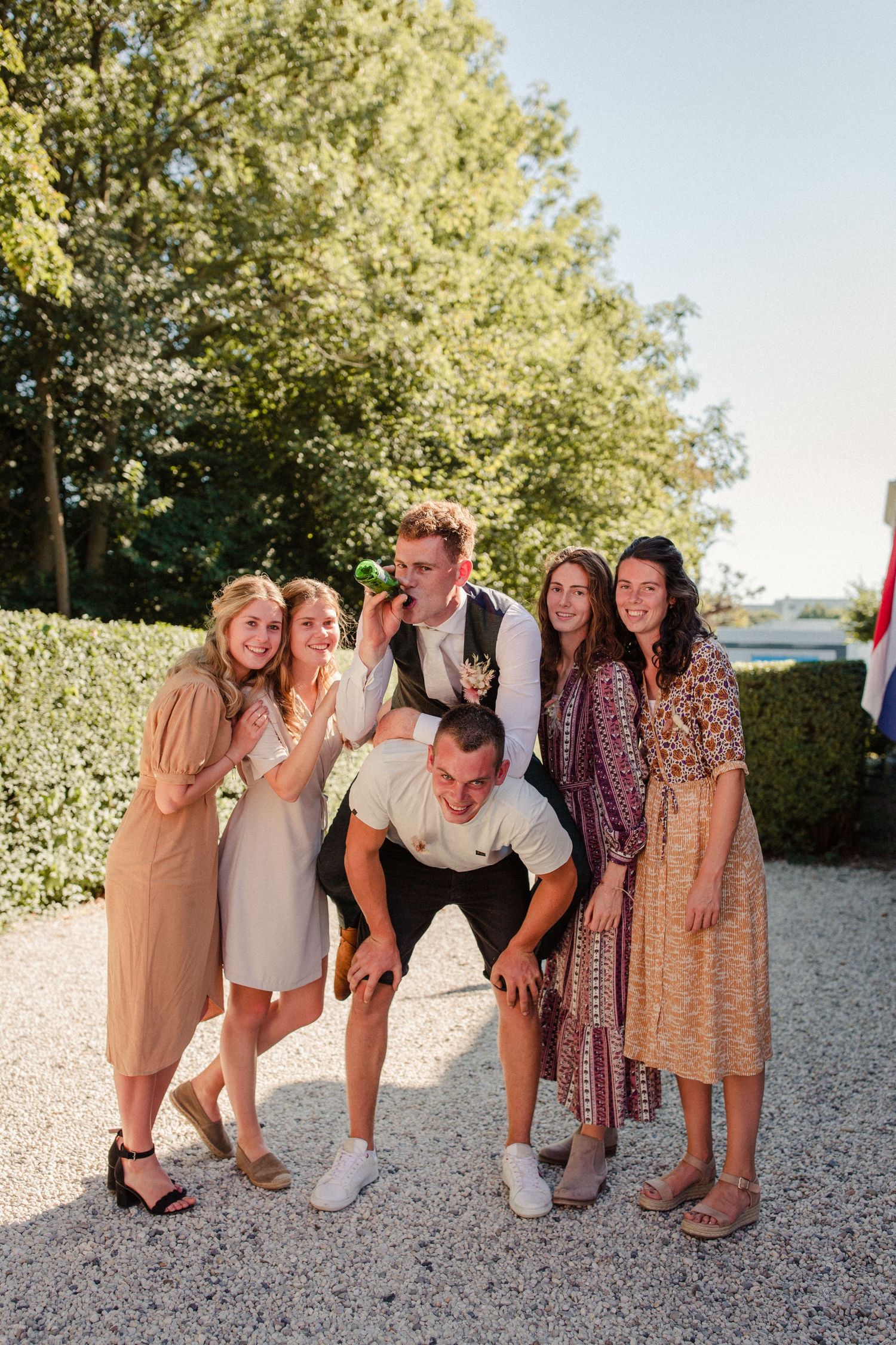 A group of people in casual and formal attire pose together on a gravel path with greenery in the background.