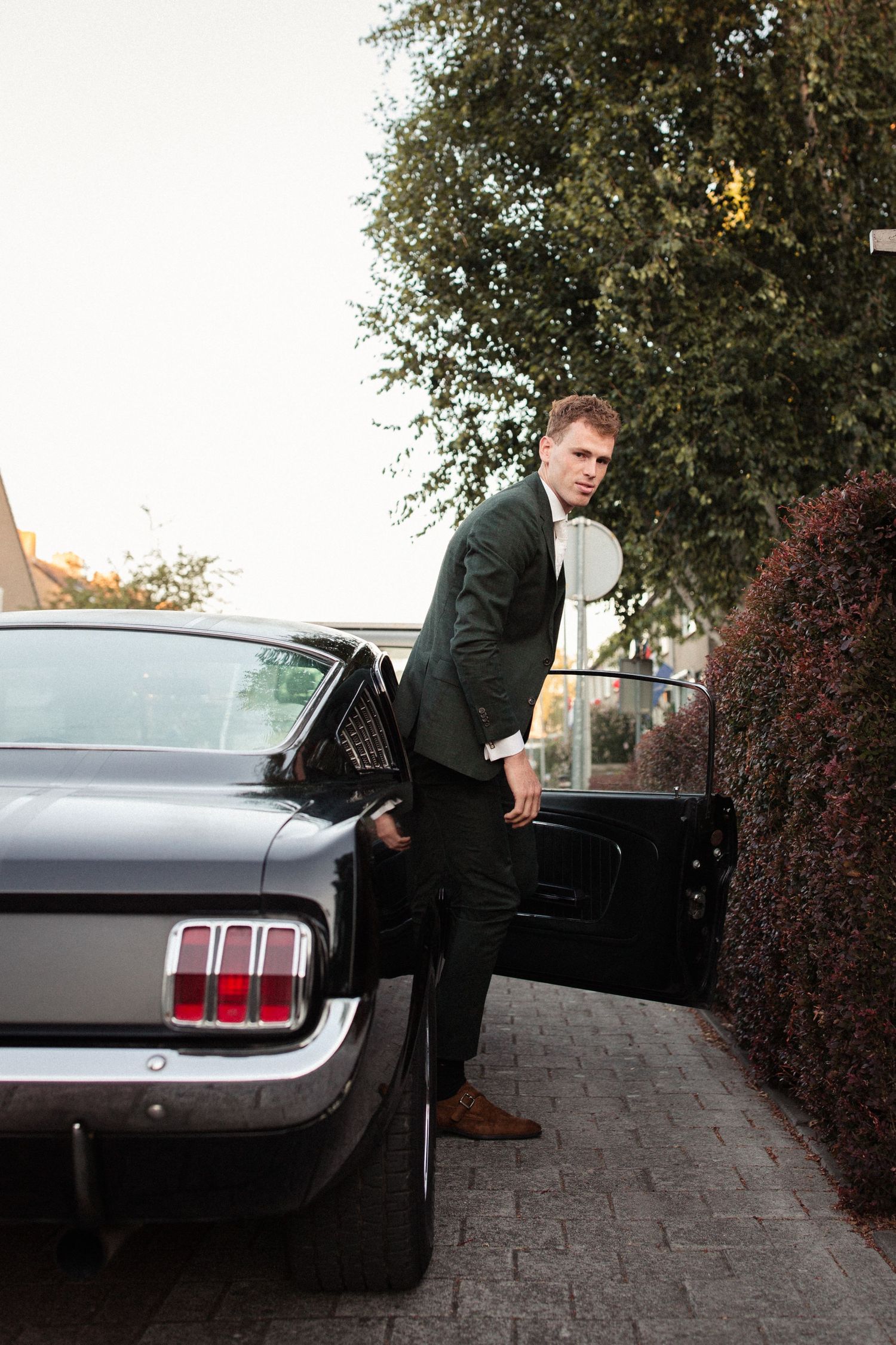 A person in a suit getting into a classic black Ford Mustang parked on a brick driveway at dusk.