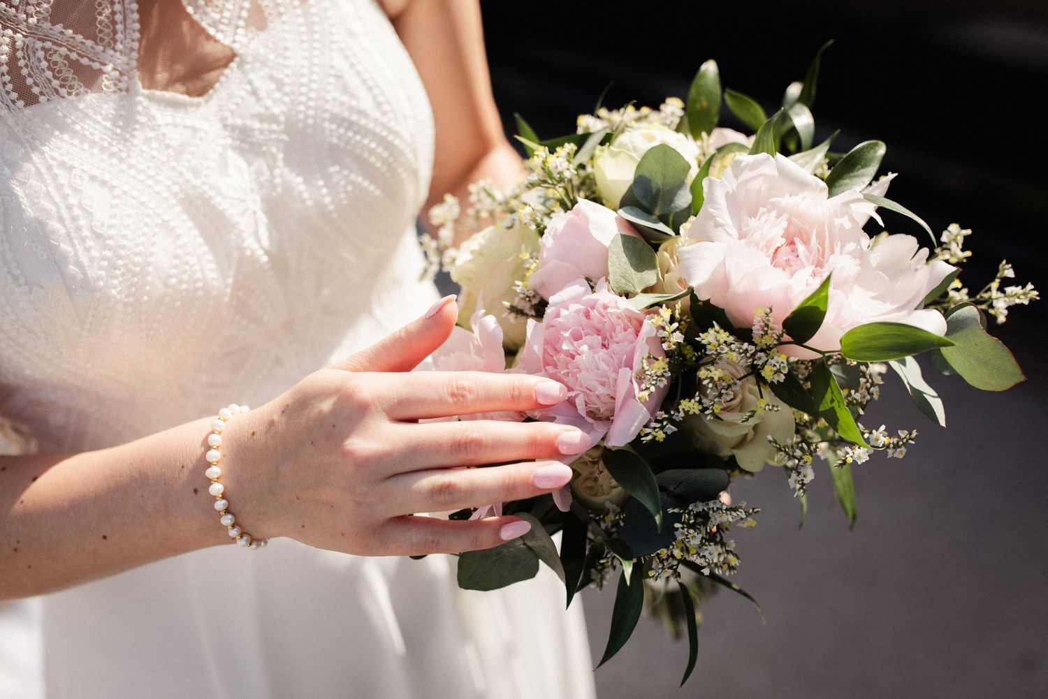 A delicate bridal bouquet with pink roses and greenery held by hands wearing a white lace dress and pearl bracelet.