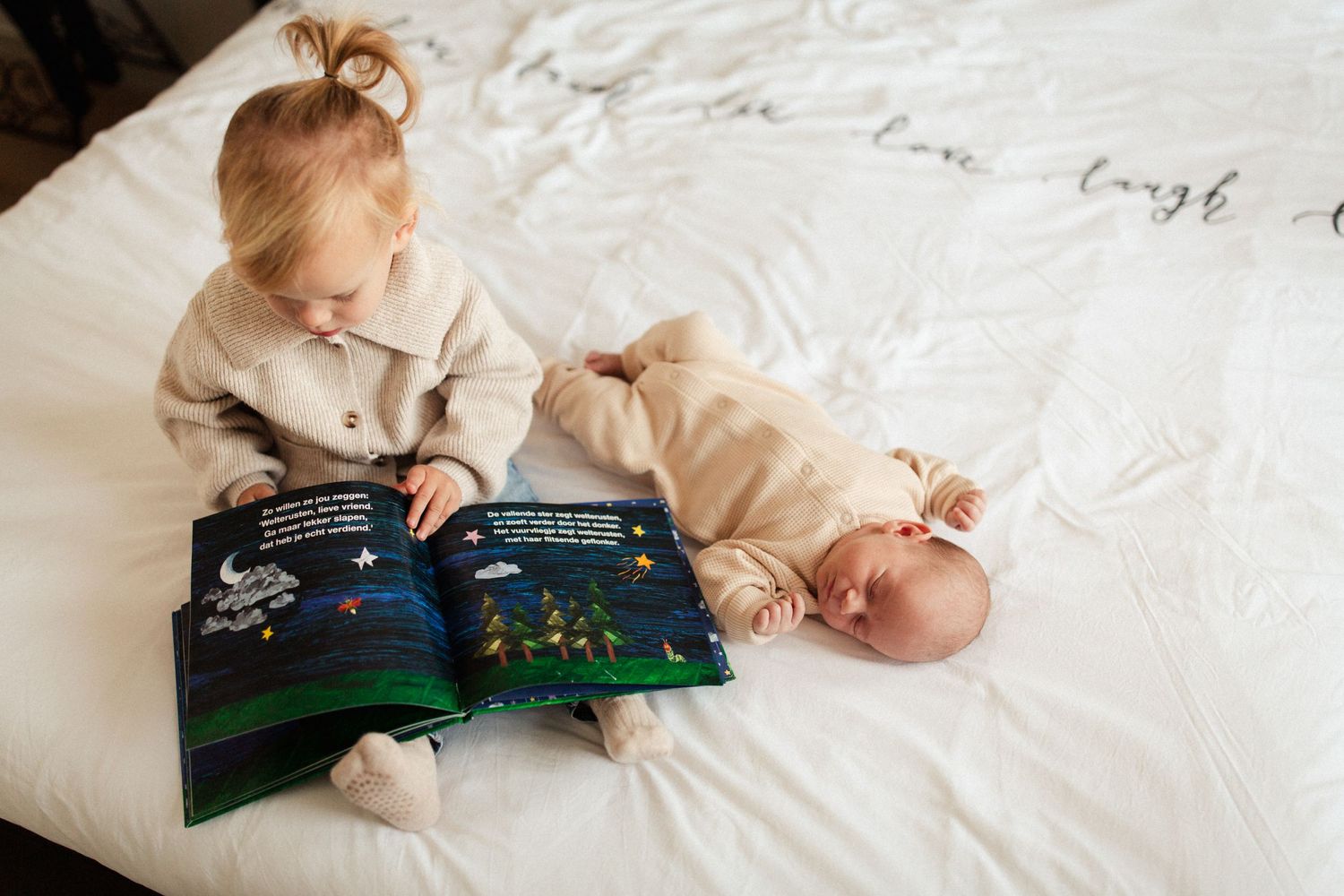 Children sharing a moment while reading a picture book together on white bedding.