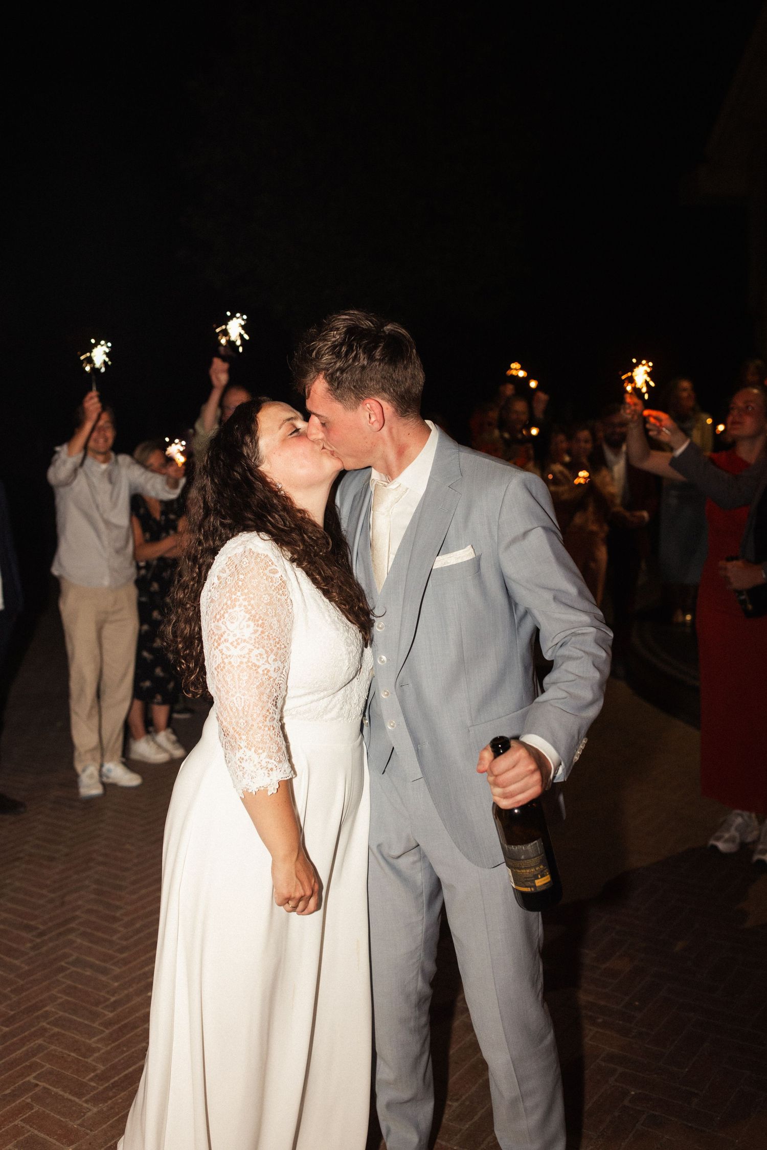 A couple shares a romantic kiss during their wedding reception at night while guests hold sparklers in the background.
