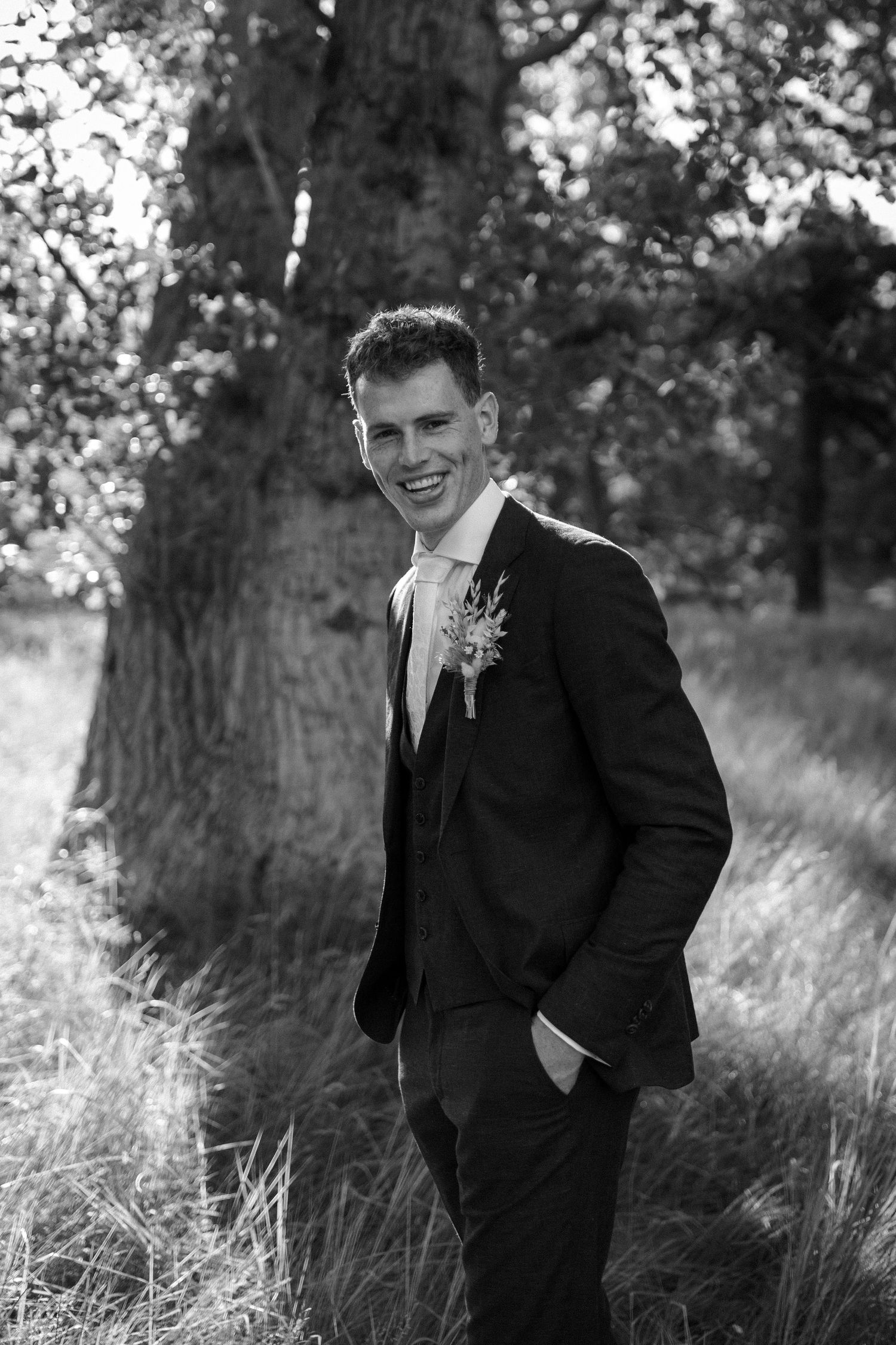 Black and white portrait of a person in a formal suit with boutonniere smiling outdoors near a tree in natural lighting.