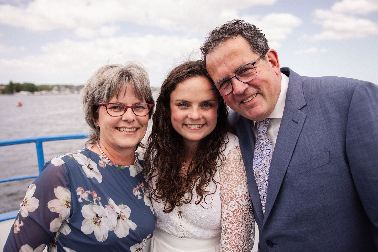 Three people smile together while standing on a boat deck with a waterway and cloudy sky in the background.