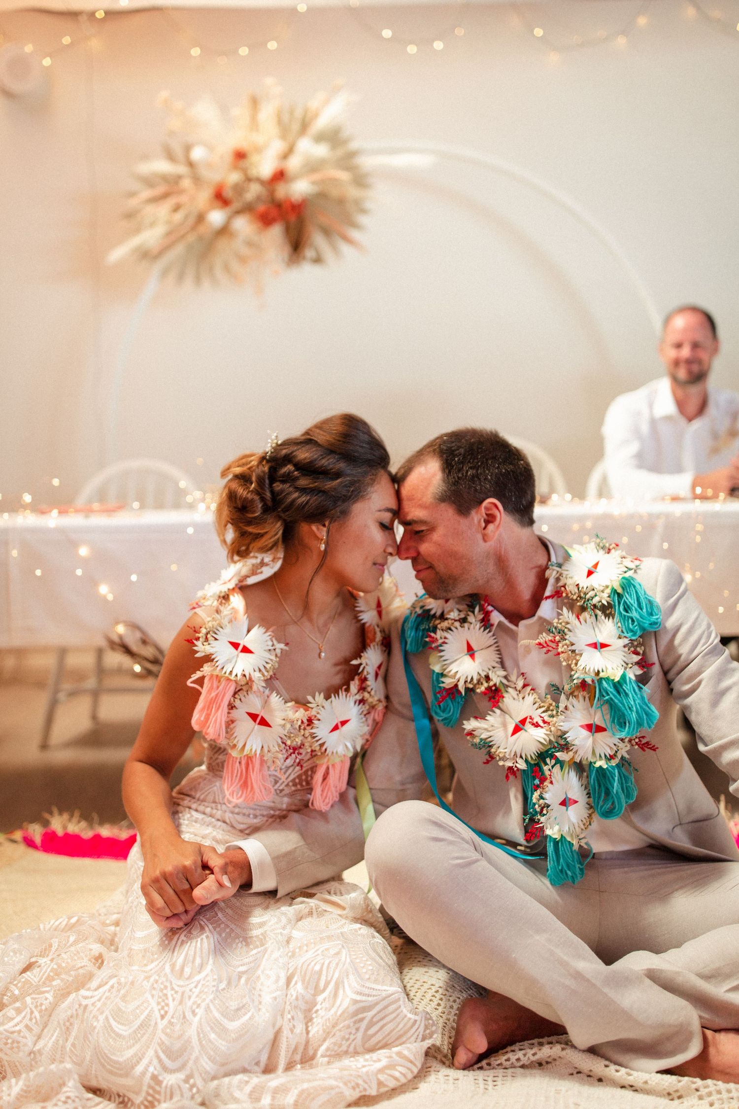 A couple in white attire share an intimate moment on the floor under decorative lighting at a celebration.