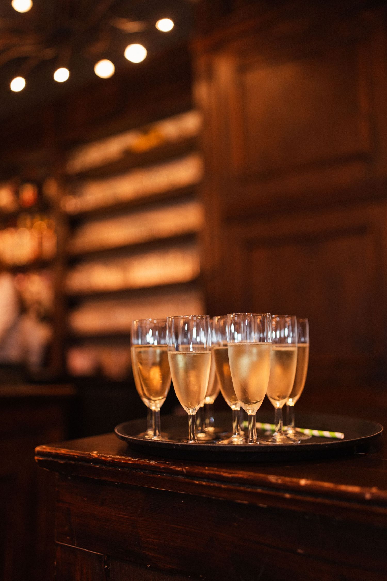 Several champagne glasses filled with golden bubbly liquid sit on a wooden tray in a dimly lit bar with bokeh lights.