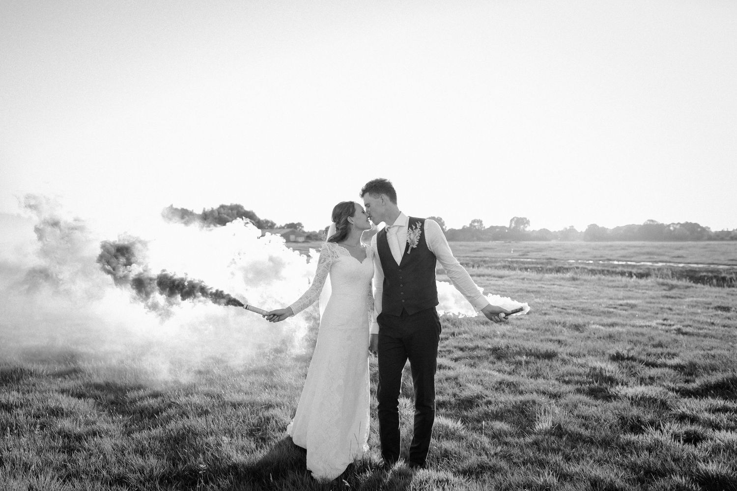 A wedding couple holding smoke flares in a misty black and white field during sunset.