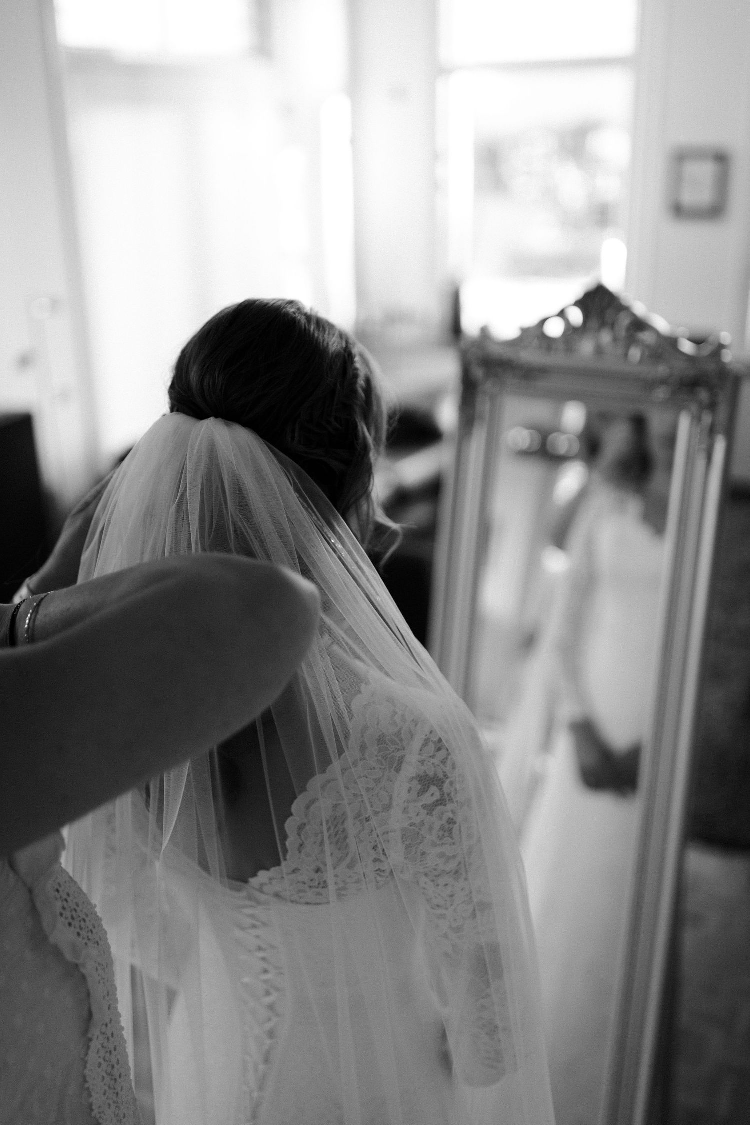A bride in a delicate lace wedding dress and veil is photographed from behind in black and white.