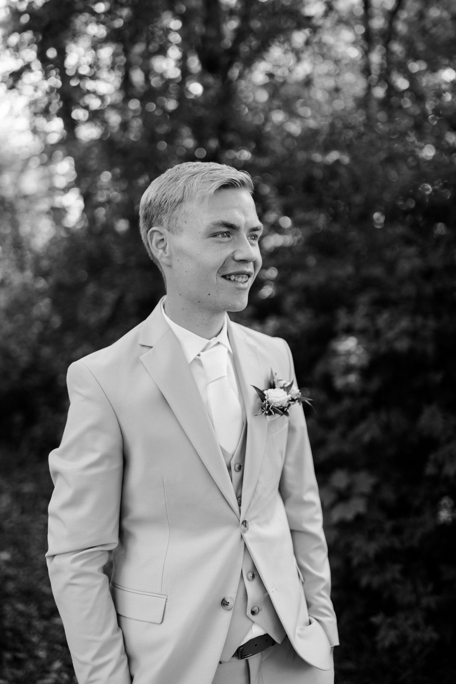 A wedding portrait in black and white showing someone wearing a light colored suit with boutonniere in an outdoor setting.