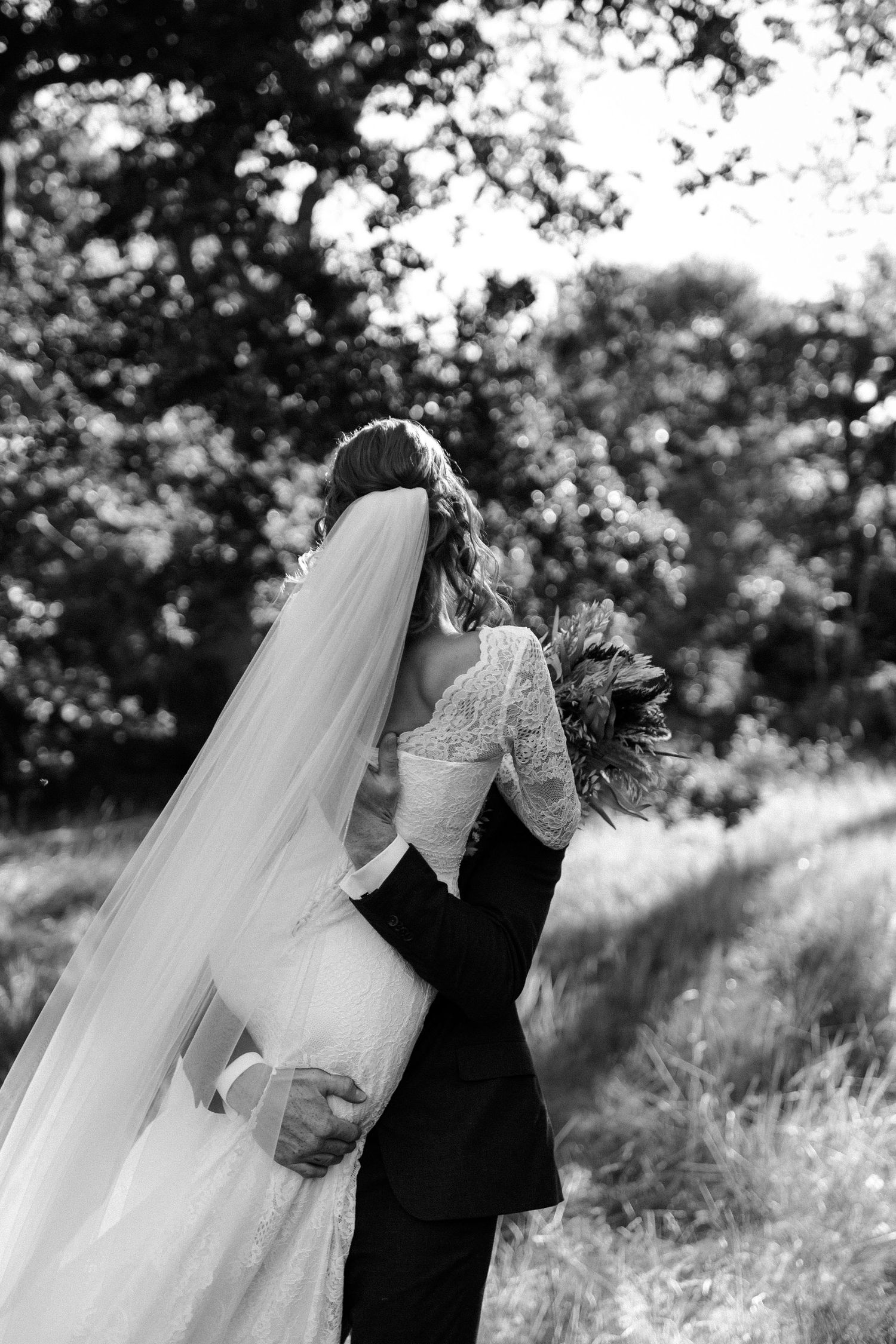 A romantic black and white wedding photo showing a couple embracing outdoors with a flowing veil in soft natural light.