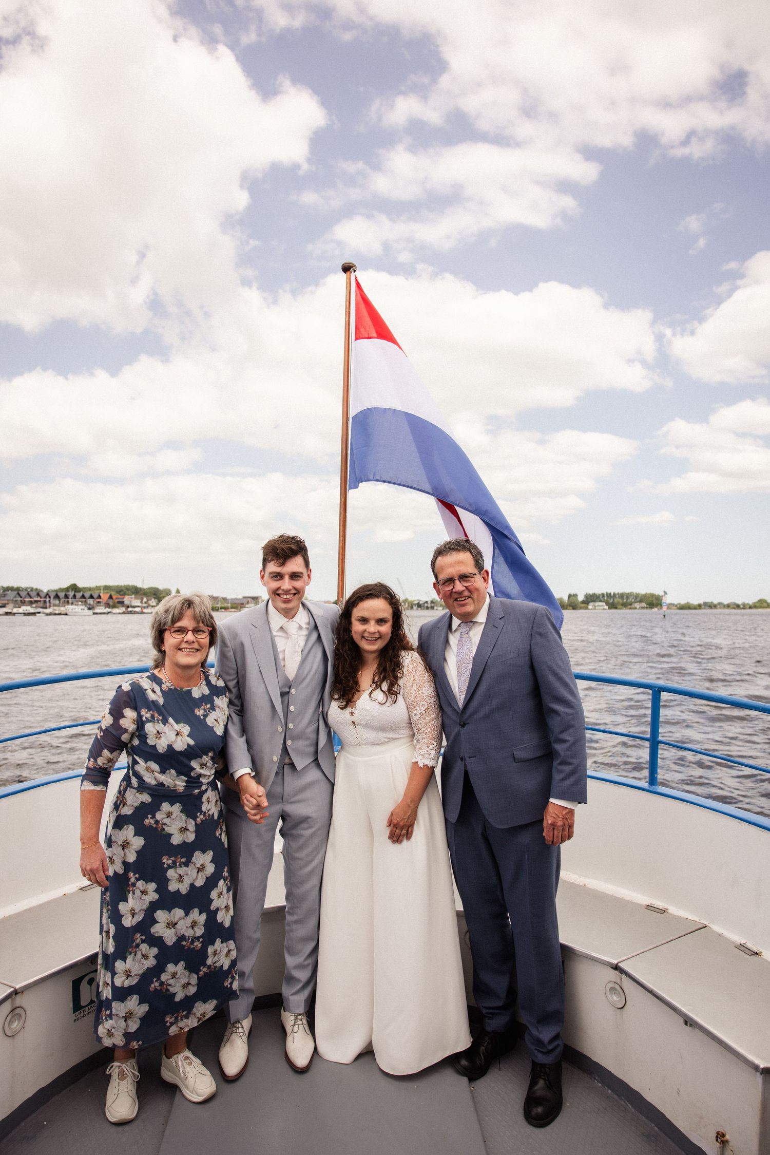 Wedding party poses on boat deck under Dutch flag with water and cloudy sky backdrop.