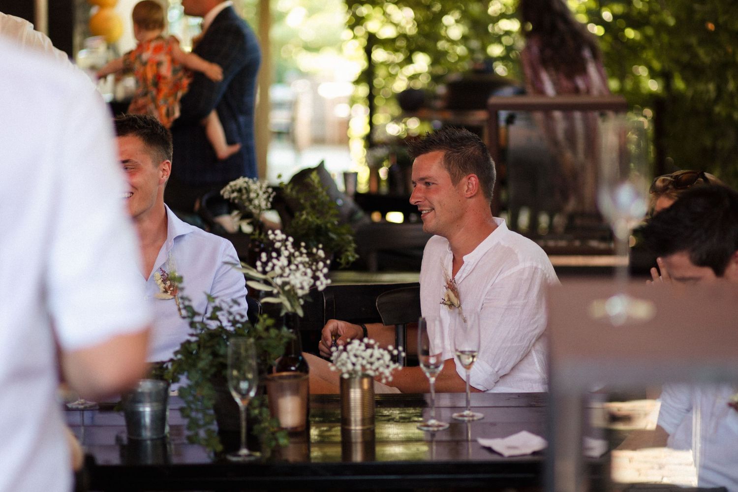People enjoying drinks at an outdoor dining table with white floral centerpieces and natural lighting.