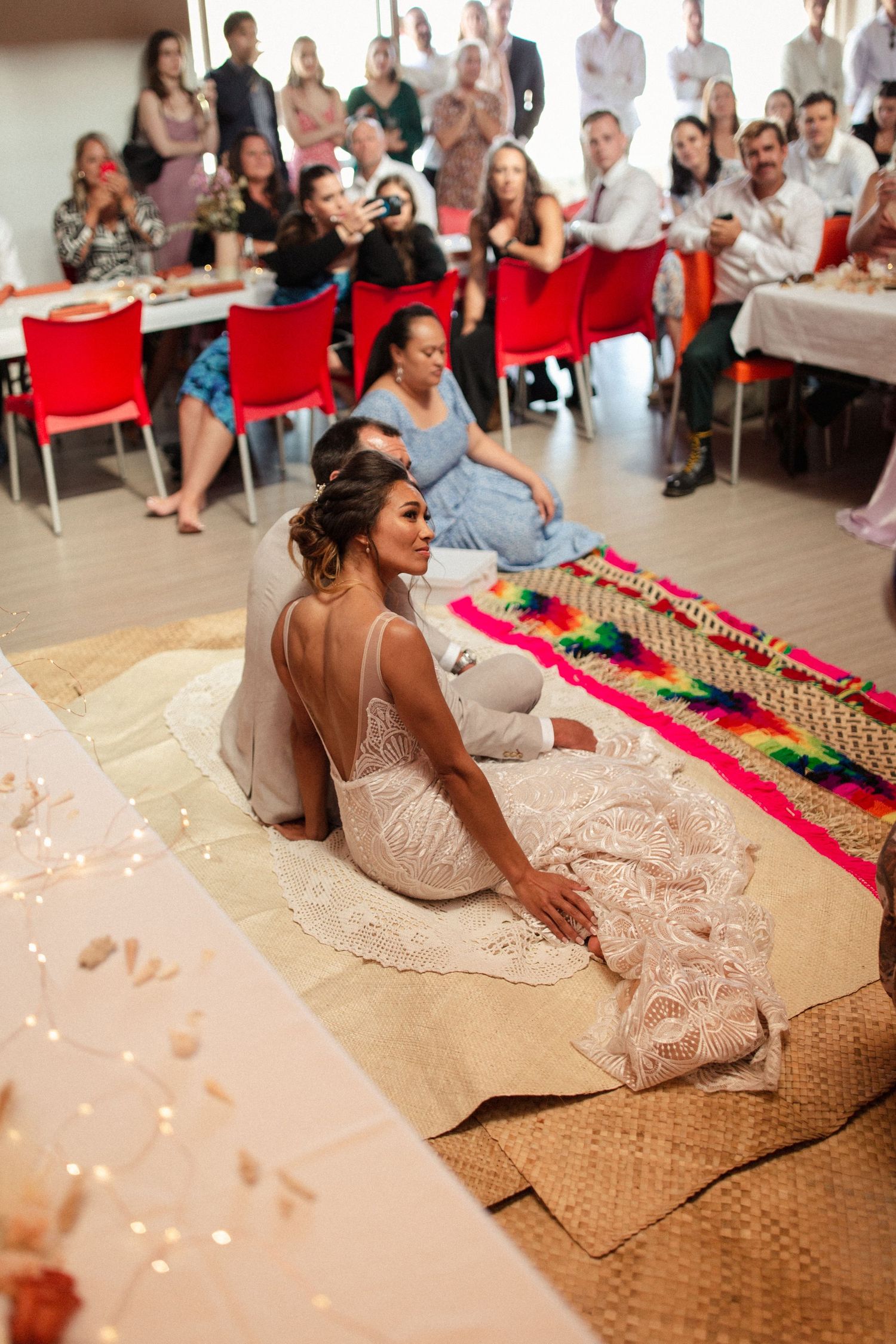 A wedding reception shows guests seated at tables with red chairs while someone sits on the floor wearing a lace dress.