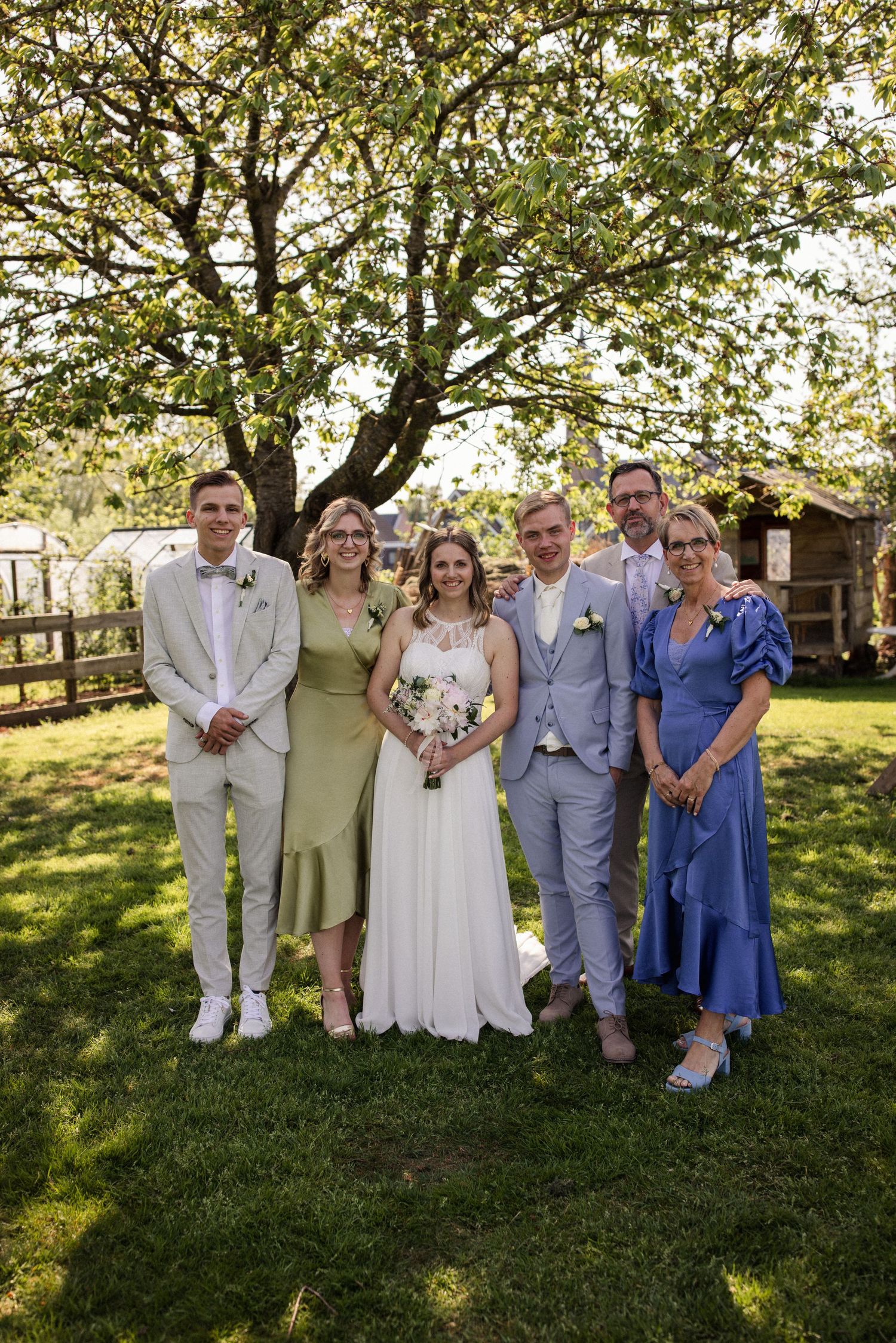 Wedding party poses together on a sunny day in a garden setting with a rustic wooden fence and tree backdrop.
