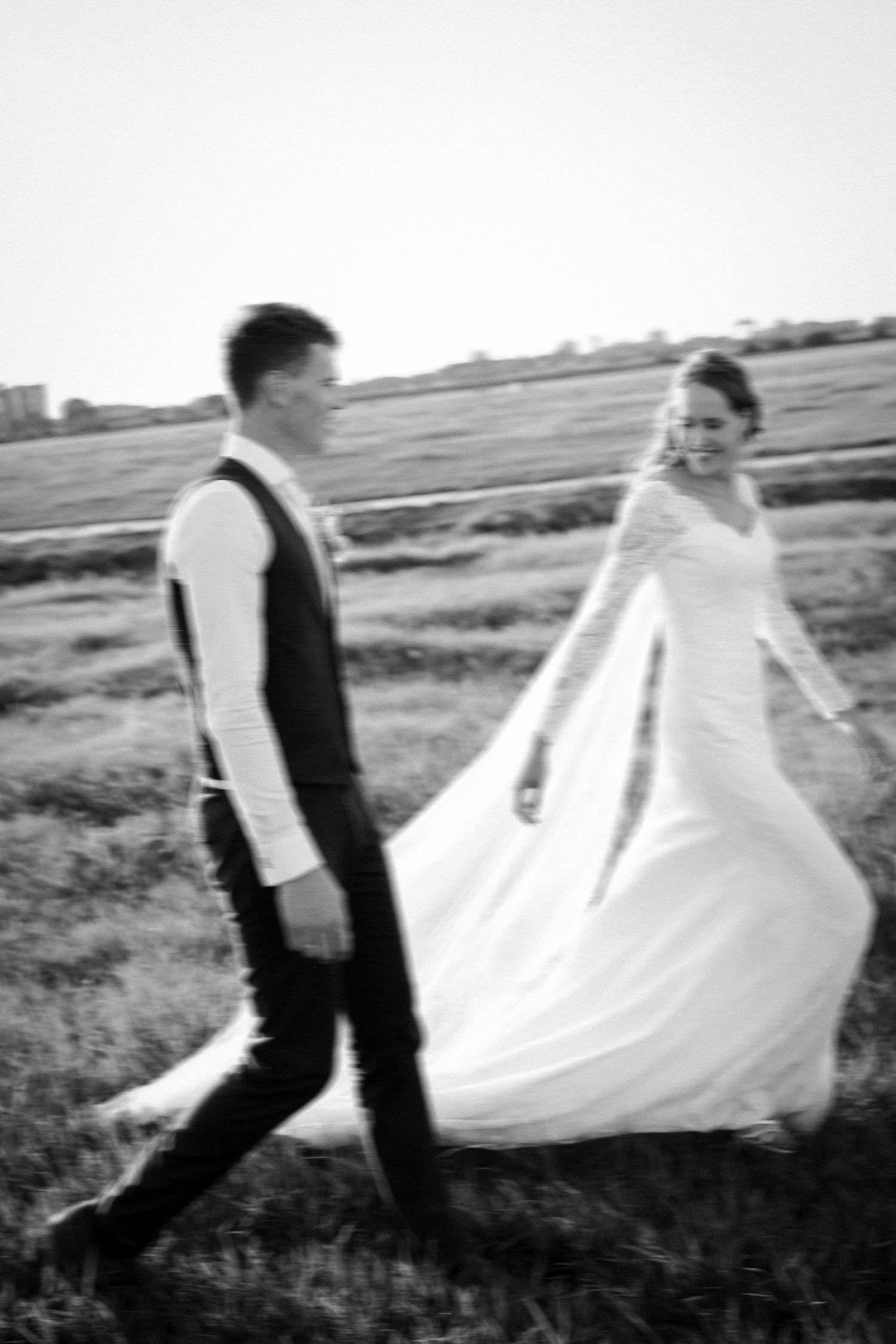 A black and white wedding photo of a couple walking in a field, with the bride's dress flowing in the wind.